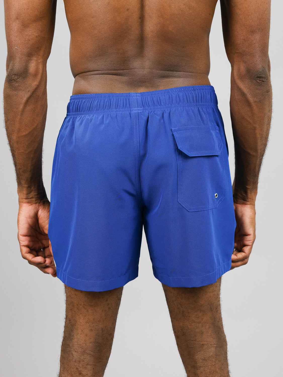 A person in Oyogu Quick Dry 4 Way Stretch Swim Trunk by ID Menswear is seen from the rear, wearing royal blue shorts made from recycled plastic bottles. The shorts have an elastic waistband and a right-side flap pocket with a button against a plain gray background.