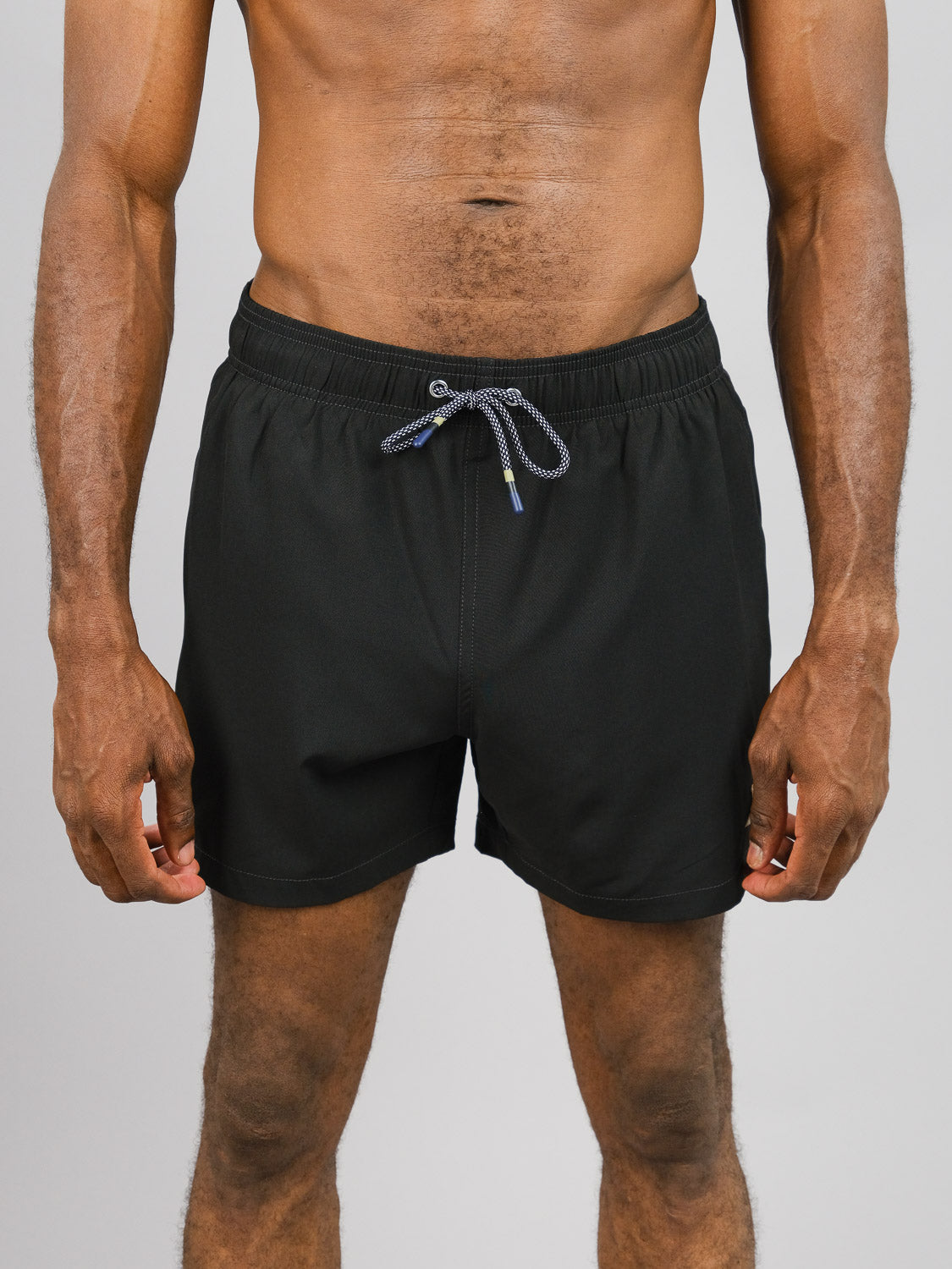 A person models ID Menswears Oyogu Quick Dry 4 Way Stretch Swim Trunk against a plain backdrop. Made from recycled plastic bottles, these black swim shorts feature a drawstring and quick-dry finish, focusing on the midsection while highlighting the legs and arms.