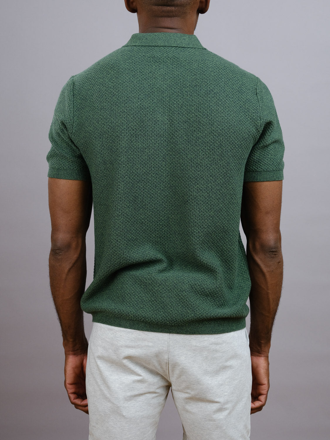 A man faces away from the camera, wearing the ID Menswear Tucson Knitted Cotton Polo in green and light gray pants—an ideal casual look set against a plain gray background.