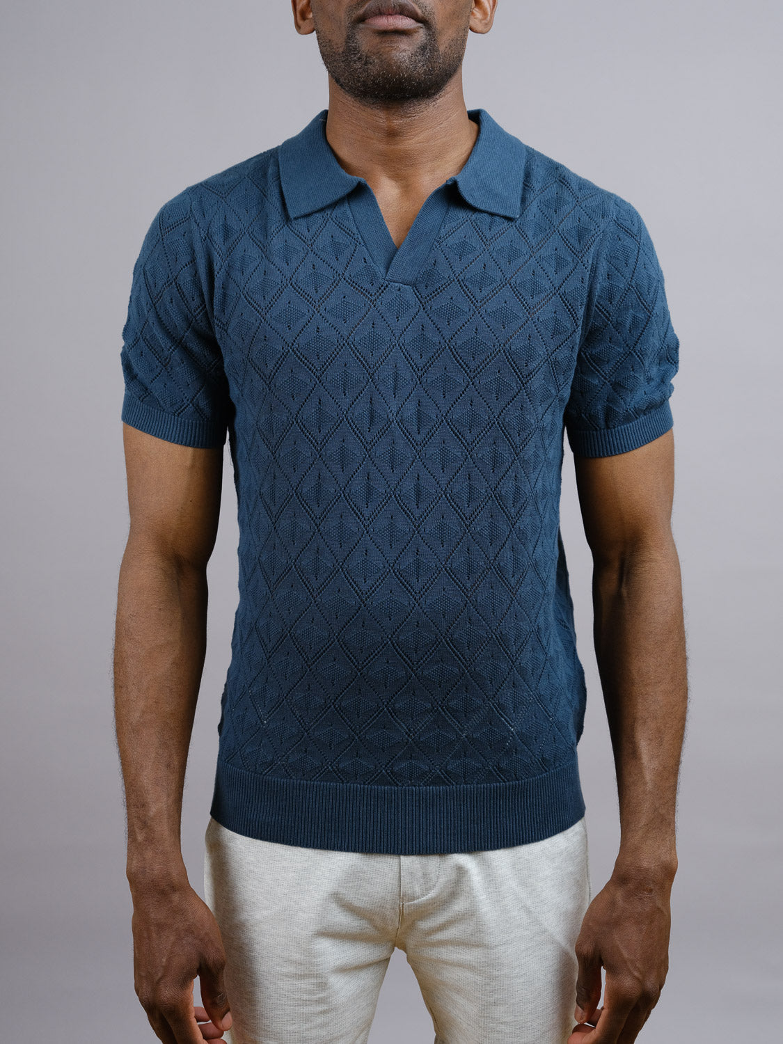 A man models the Agra Knitted Cotton Polo by ID Menswear—a textured blue, collared top—paired with light pants against a gray backdrop, highlighting its breathable comfort and laid-back style.