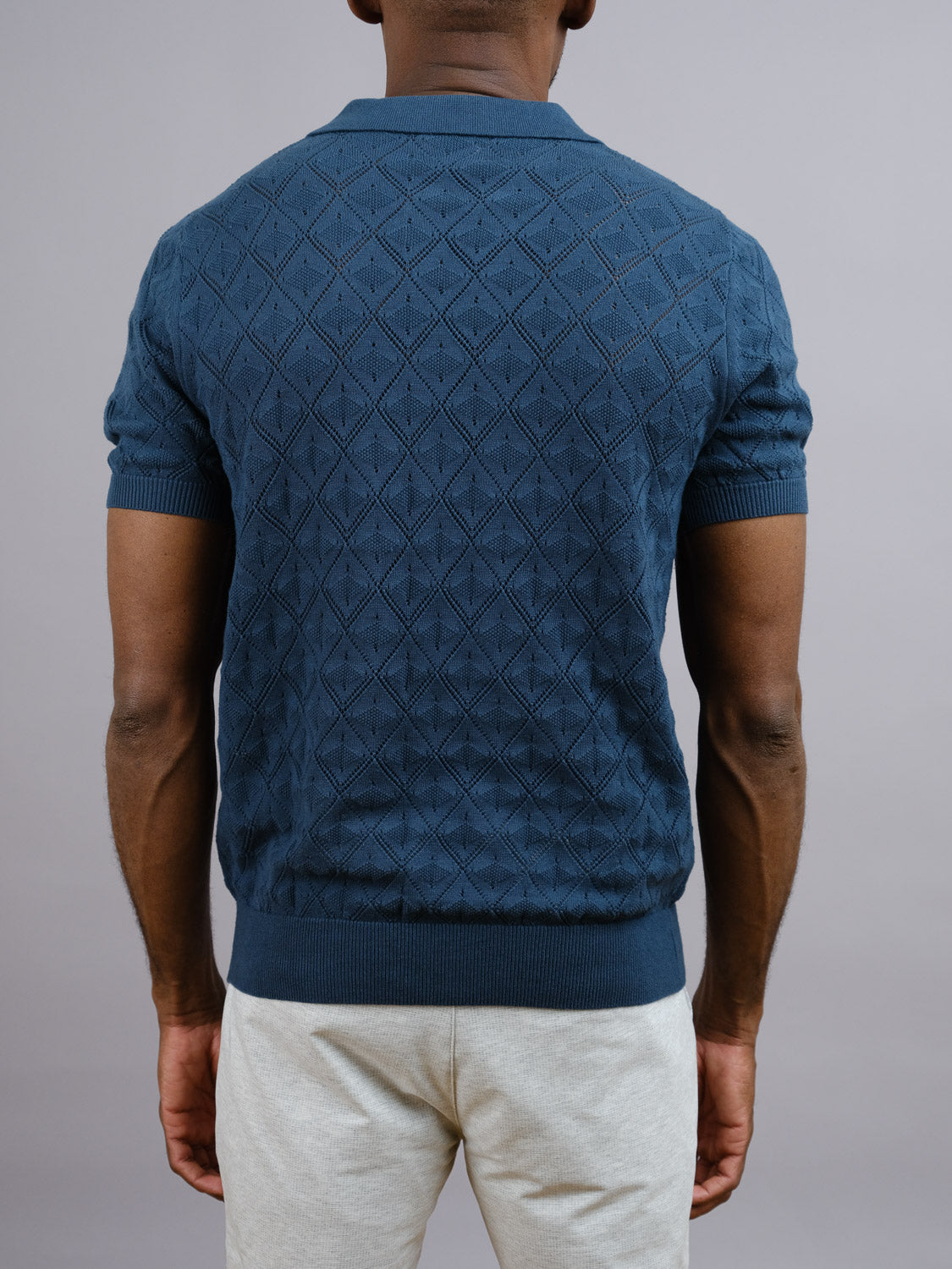 A man faces away, wearing the Agra Knitted Cotton Polo by ID Menswear—a textured short-sleeve navy blue sweater with a diamond pattern—paired with light-colored pants, ideal for casual wear, set against a plain gray background.