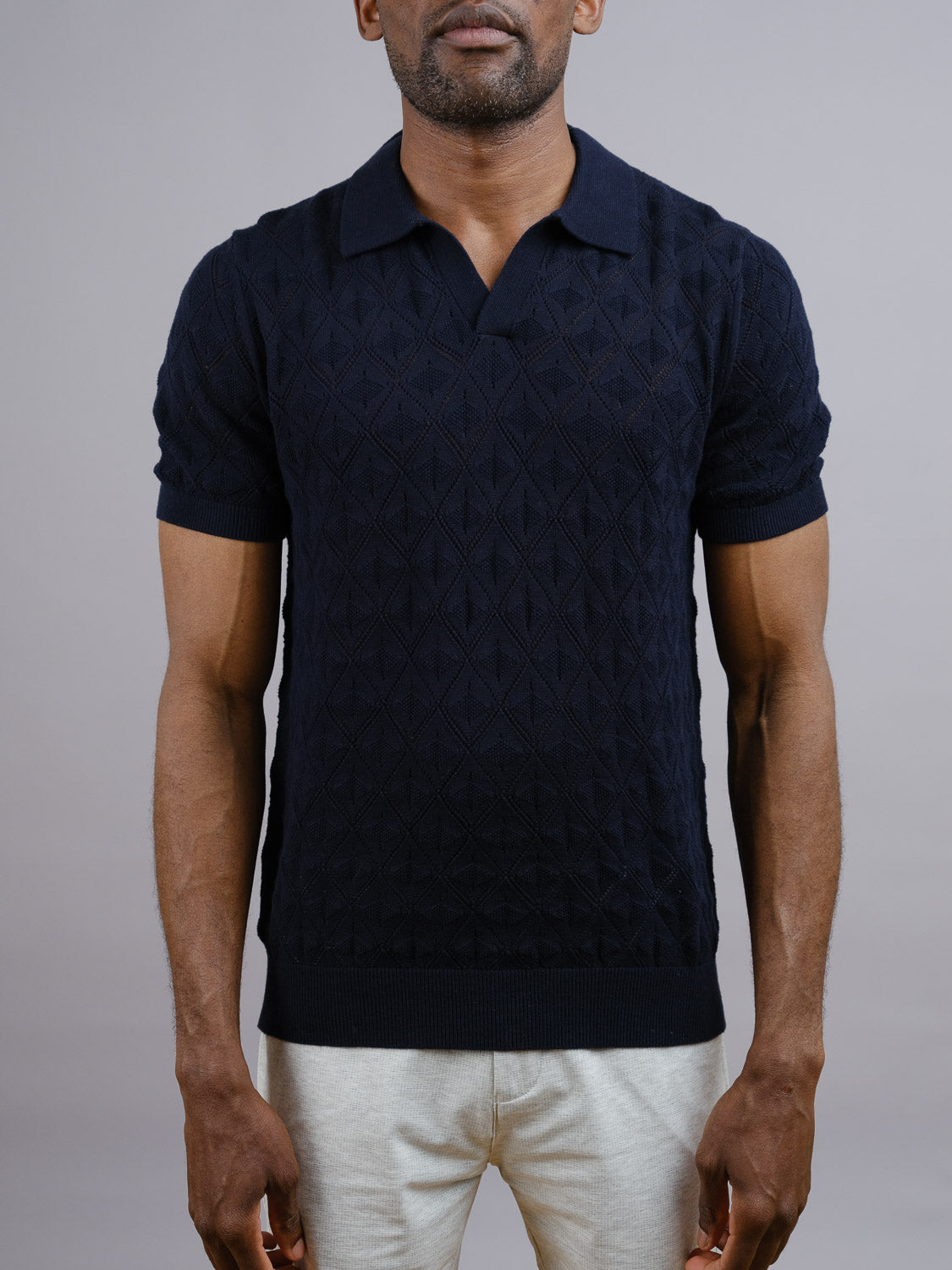 A man wears the Agra Knitted Cotton Polo by ID Menswear—a short-sleeve, dark navy cotton-knit shirt with a diamond pattern and collar—paired with light pants, set against a plain gray background for refined casual style.