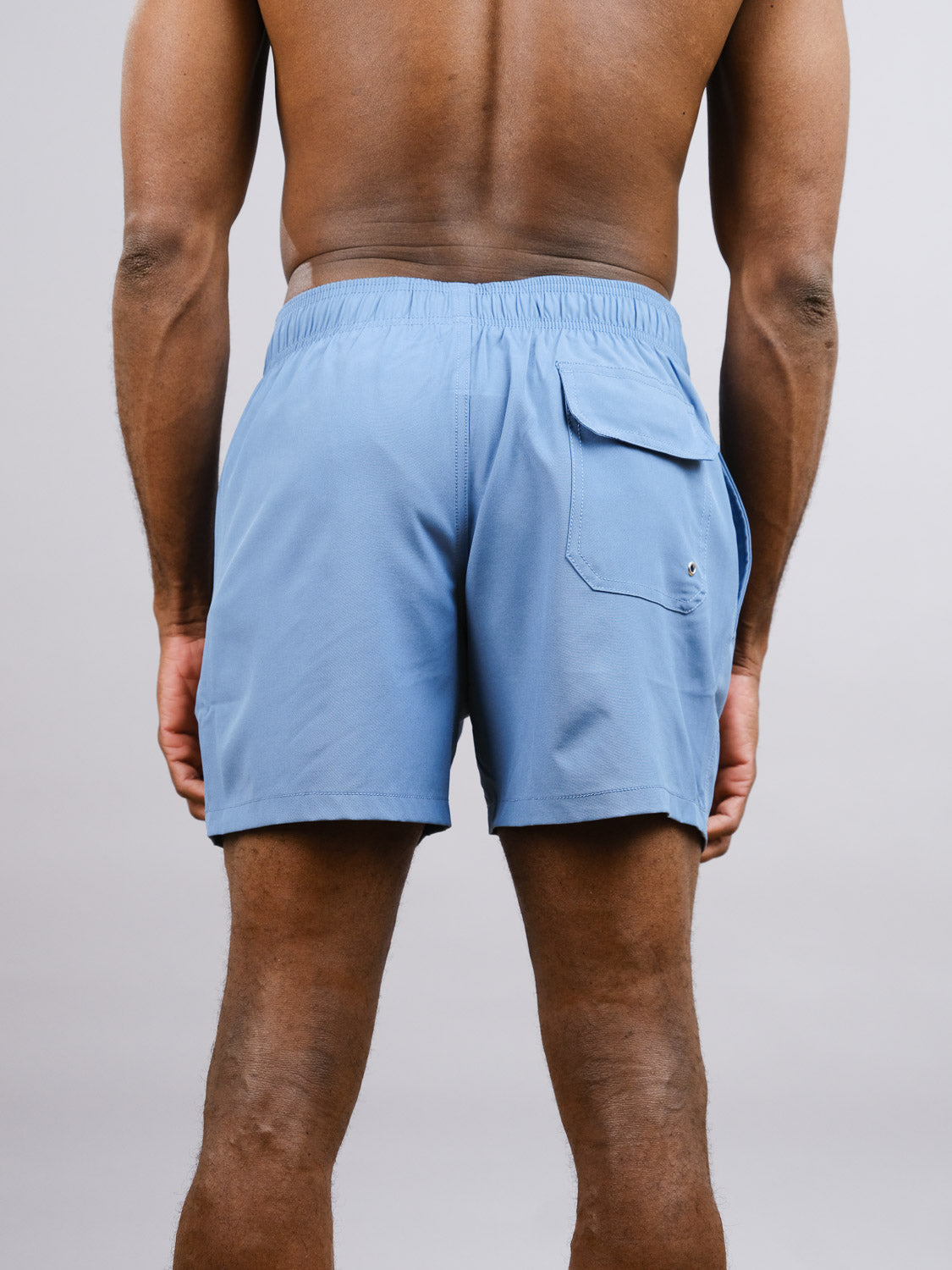 A man is seen from the back in a plain light gray setting, wearing ID Menswears Oyogu Quick Dry 4 Way Stretch Swim Trunk in light blue with an elastic waistband and right-side flap pocket, made from recycled plastic bottles.