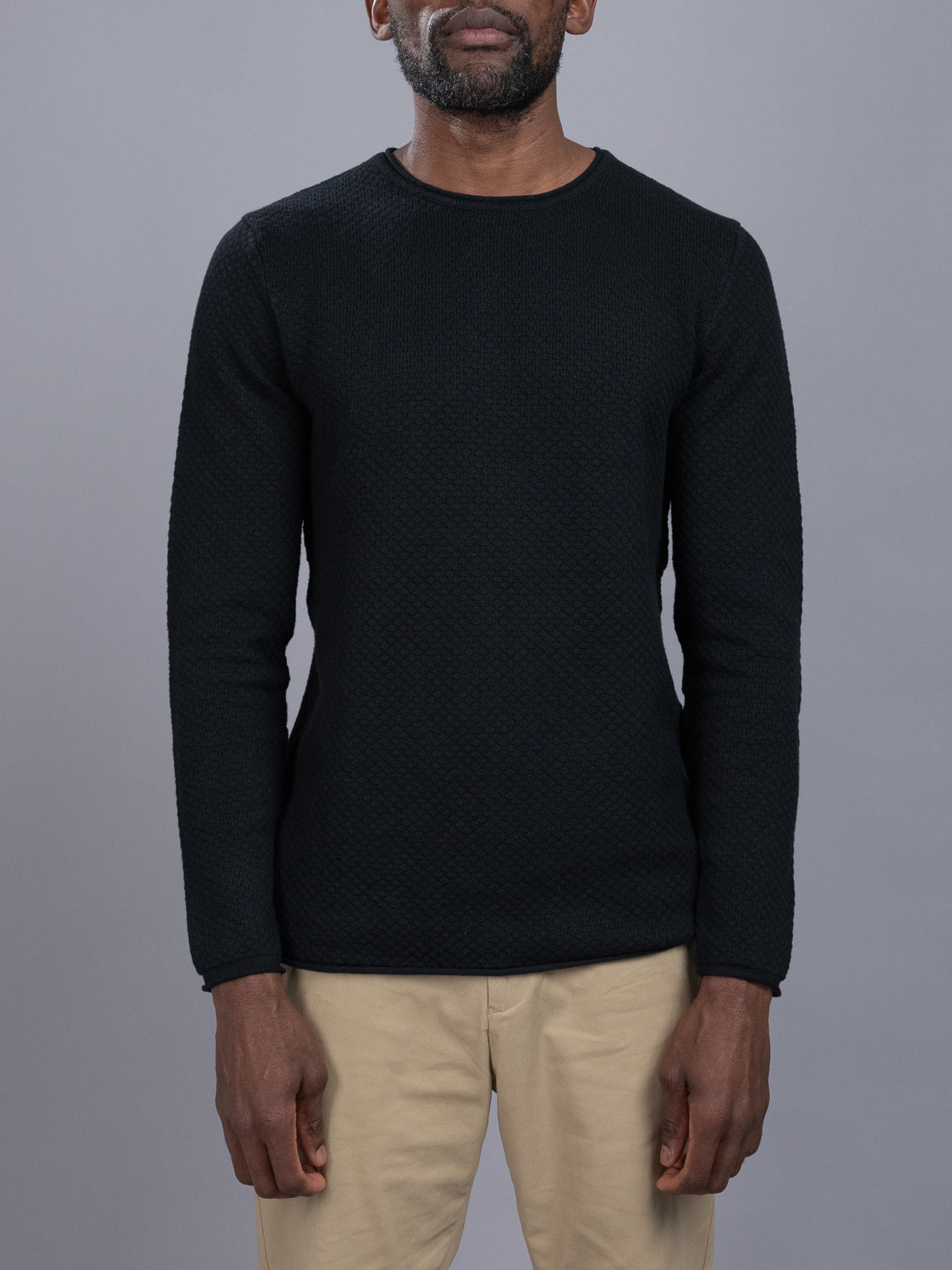 An individual showcases the Westside Rolled Crew Neck Sweater from ID Menswears exclusive collection, paired with beige pants against a gray backdrop. The black textured long-sleeve sweater seamlessly combines style and comfort, while the models face is obscured.