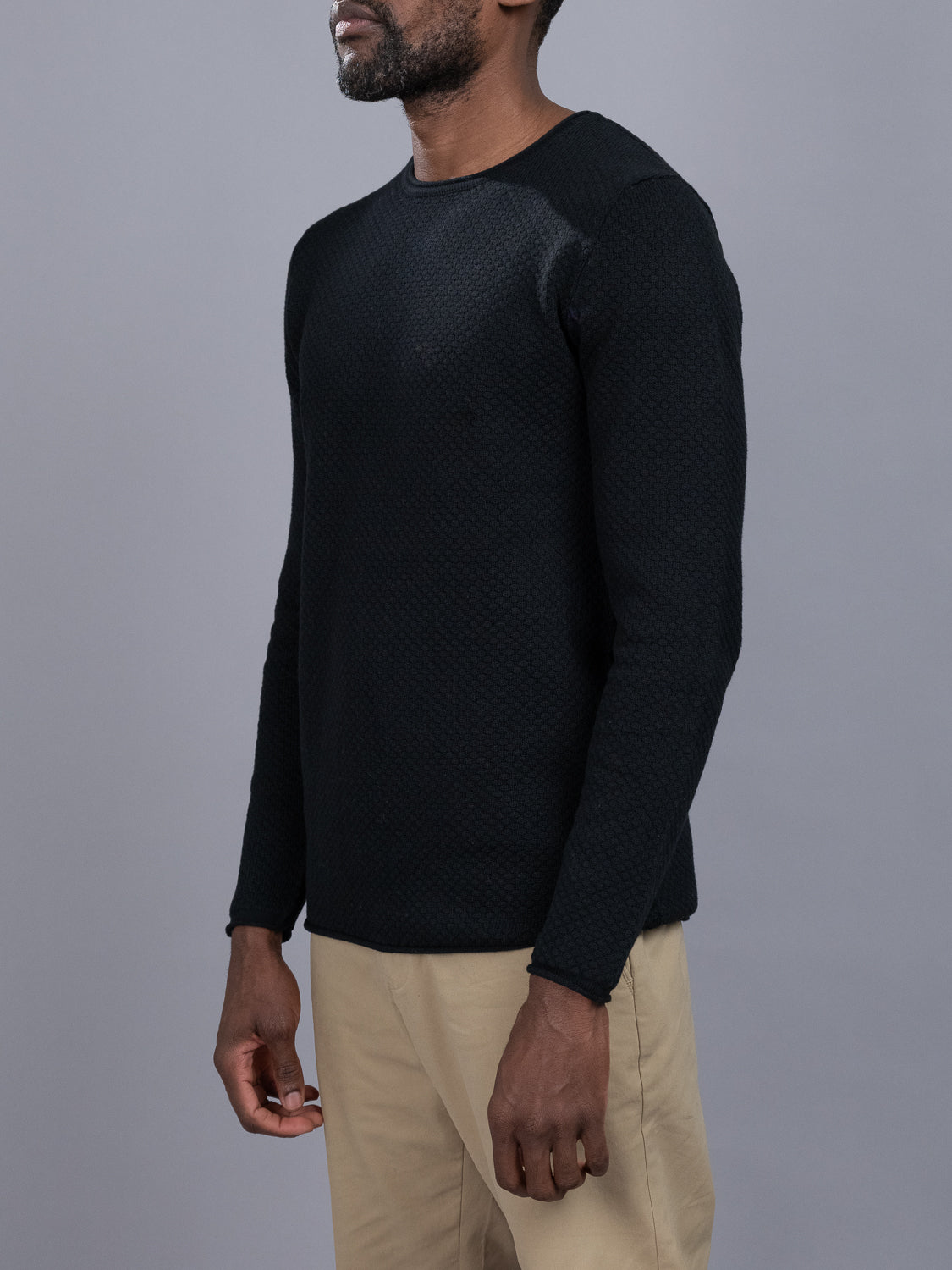A person in a textured black Westside Rolled Crew Neck Sweater from ID Menswear and beige pants stands against a gray background. The photo captures them from shoulders to thighs, facing slightly right, highlighting an elegant piece from the new knitwear collection.