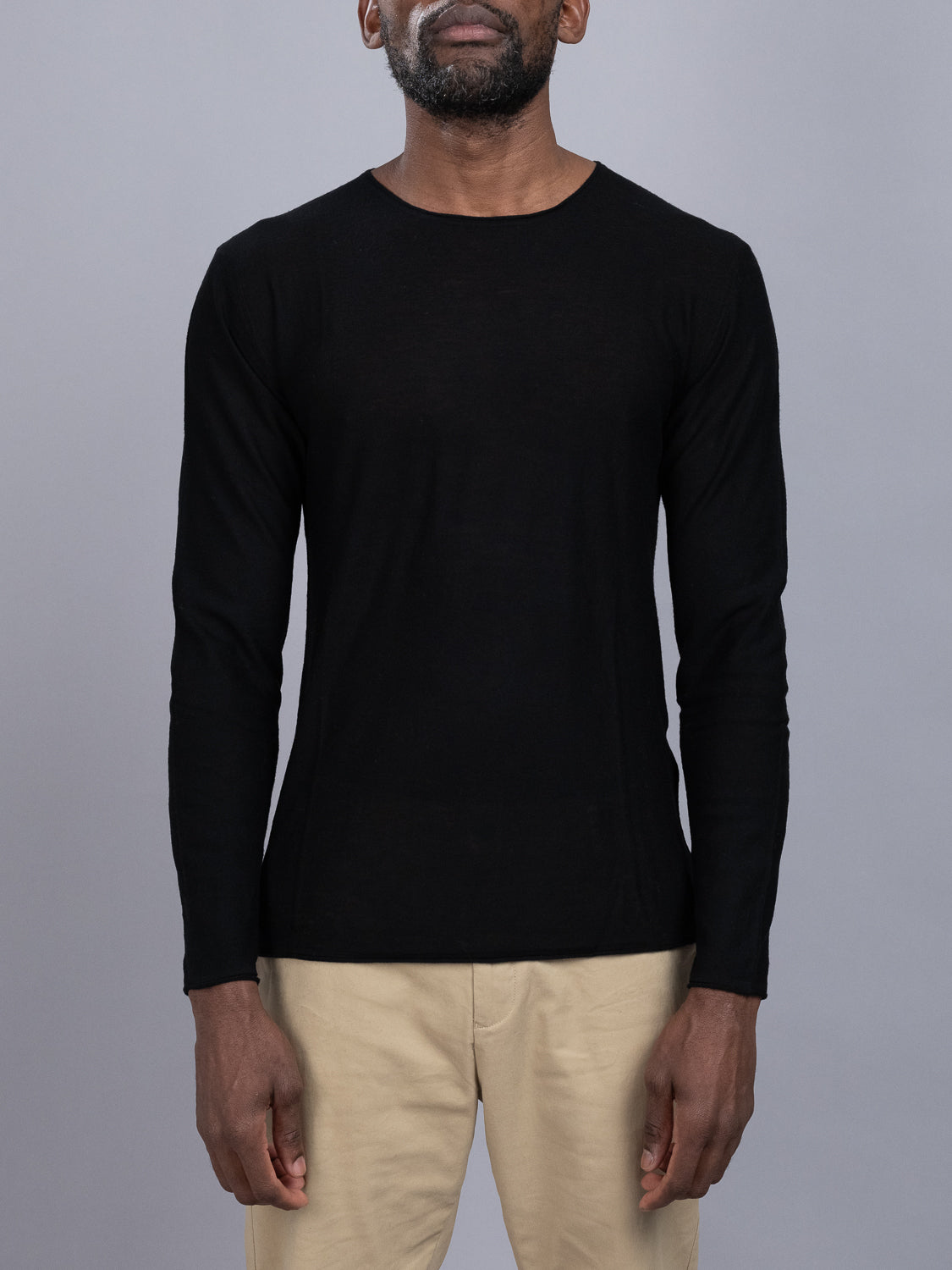 A person stands against a gray background, wearing an ID Menswear Vogue Lightweight Merino Sweater in black and beige pants, with their arms relaxed by their sides.