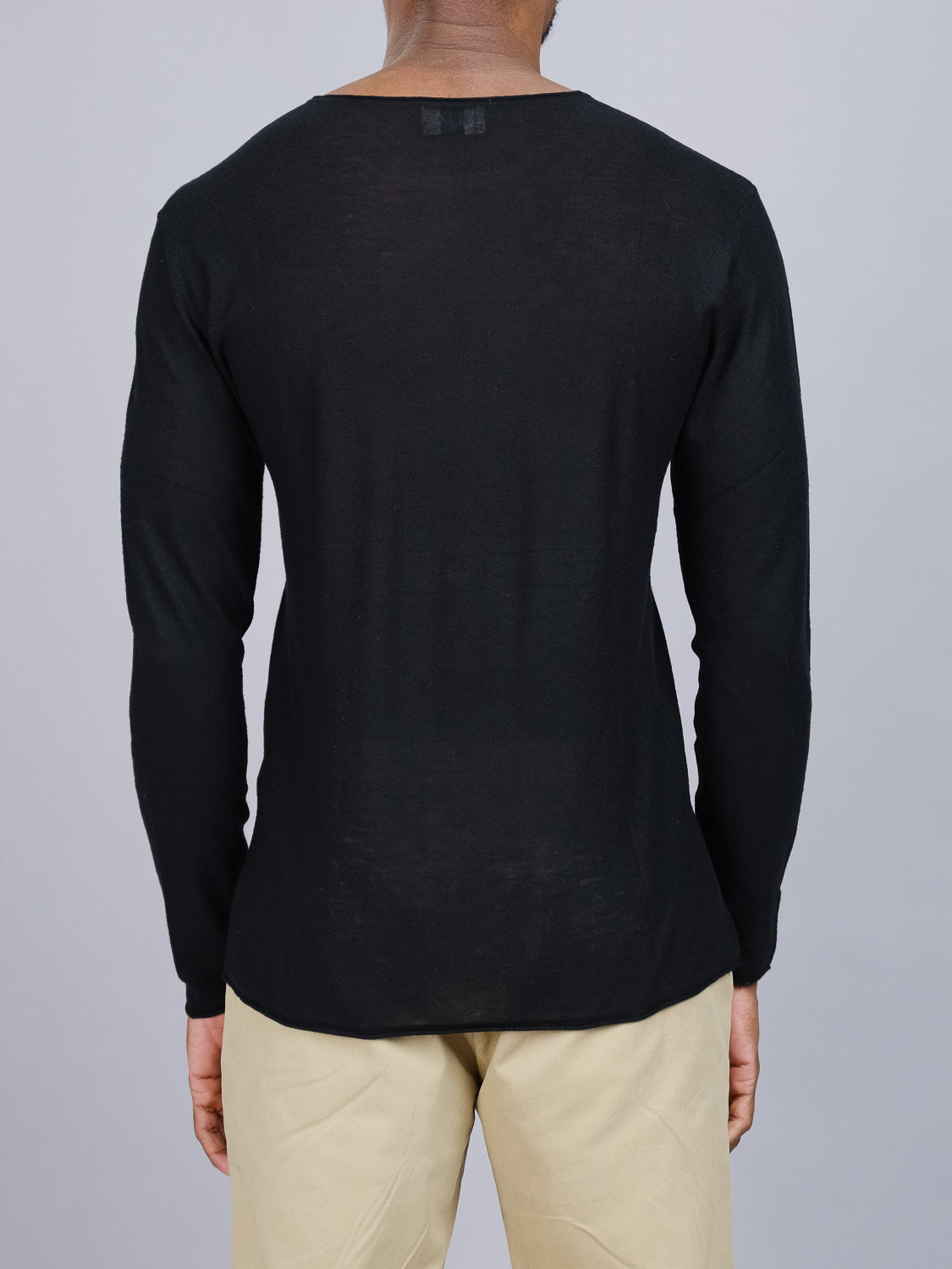 A person wearing an ID Menswear Vogue Lightweight Merino Sweater and beige pants stands with their back to the camera against a plain gray background.