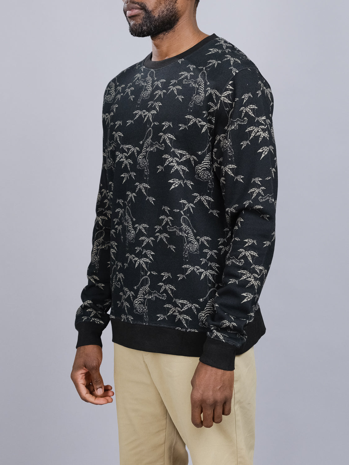 A person is styled in a black Tiger Bamboo Printed Sweatshirt by ID Menswear, showcasing an intricate tiger and foliage motif. The round-necked, long-sleeved sweatshirt pairs well with beige pants against a plain light gray backdrop.