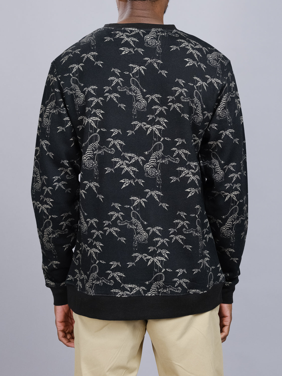 The individual wears an ID Menswear Tiger Bamboo Printed Sweatshirt made from organic cotton, adorned with detailed tigers and bamboo motifs. They face away from the camera, paired with beige pants against a plain gray background.