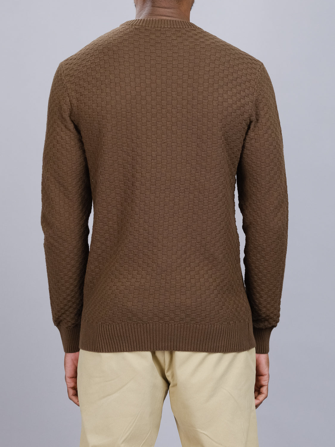 Wearing the ID Menswear Turku Cotton Sweater and beige pants, a person stands against a gray background. The brown checked knit pattern of this regular-fit sweater enhances its charm.