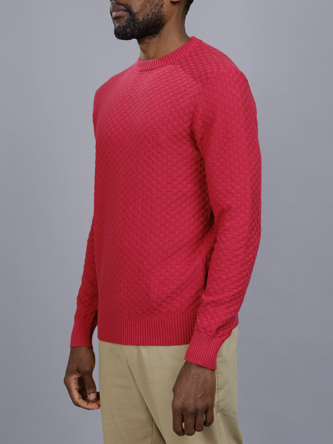 A person wearing the Turku Cotton Sweater by ID Menswear, featuring a stylish knit pattern in regular fit, and beige pants stands against a gray background. The checked red sweater showcases its subtle design as the person faces sideways to highlight its texture and form.