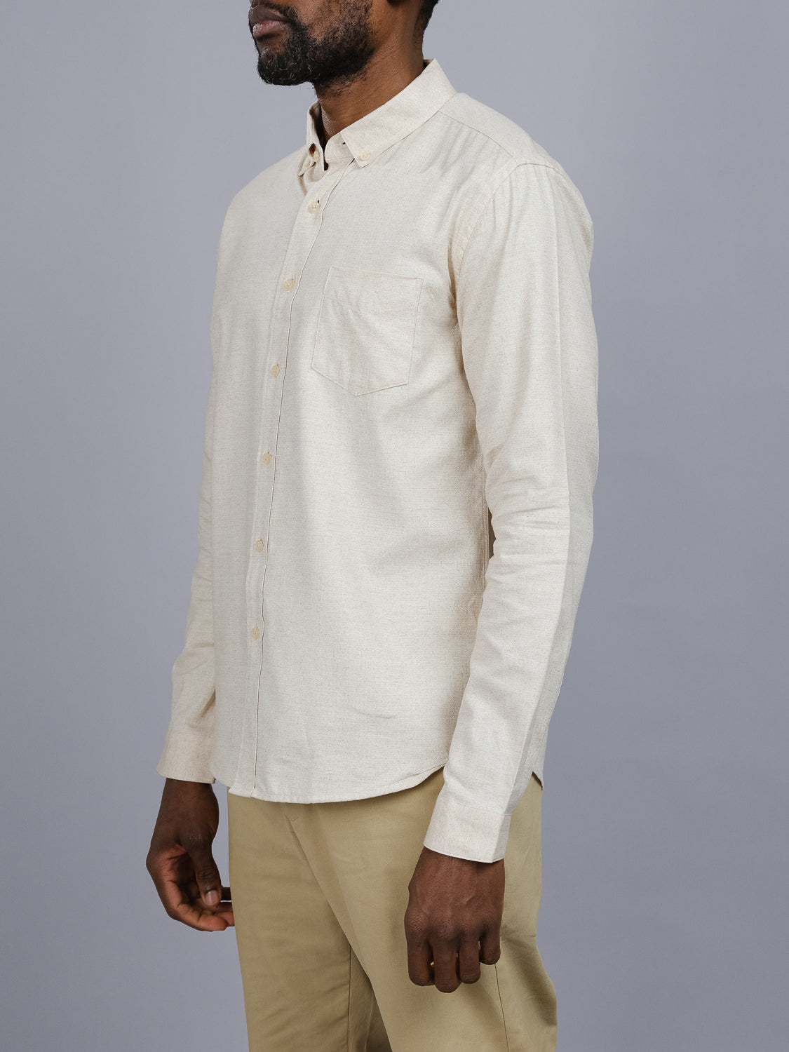A person wearing the ID Menswear Maya Fine Herringbone Shirt stands against a gray background, facing slightly left. The beige flannel shirt features a pocket and is paired with beige pants, embodying a relaxed yet refined style. Only the persons torso and part of their face are visible.