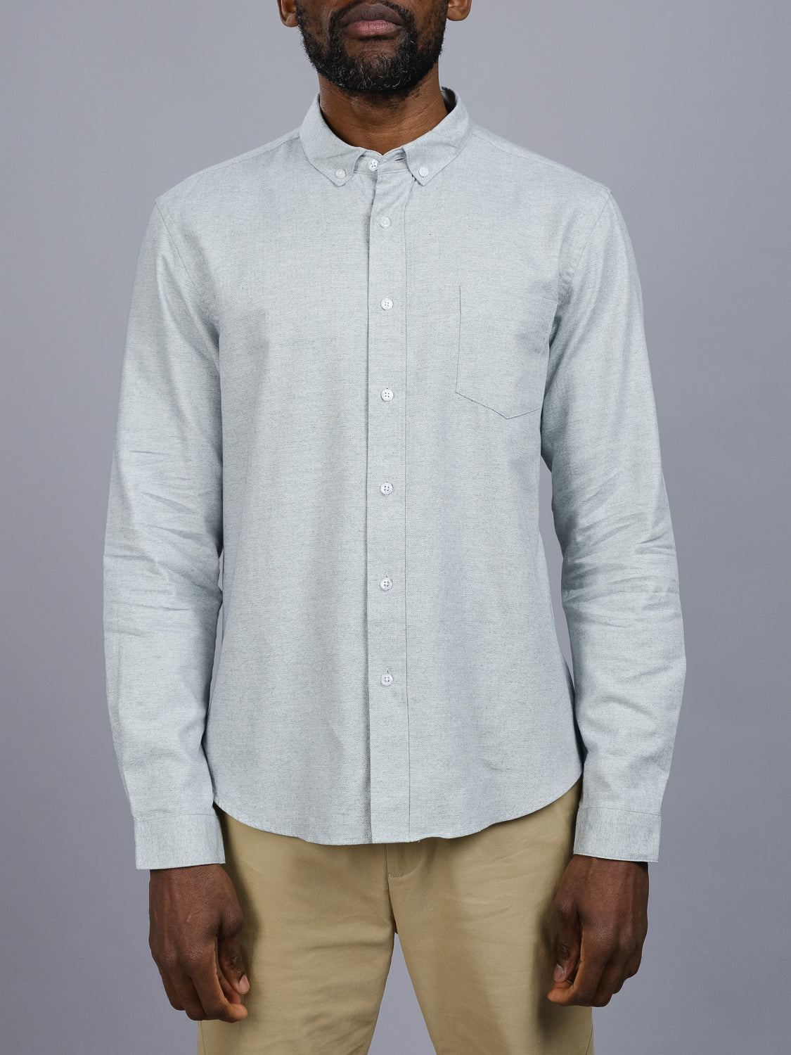 A person wears the ID Menswear Maya Fine Herringbone Shirt in light gray, crafted from 100% cotton with a left chest pocket, paired with beige pants. The scene is set against a gray background, and the subjects head is partially visible.