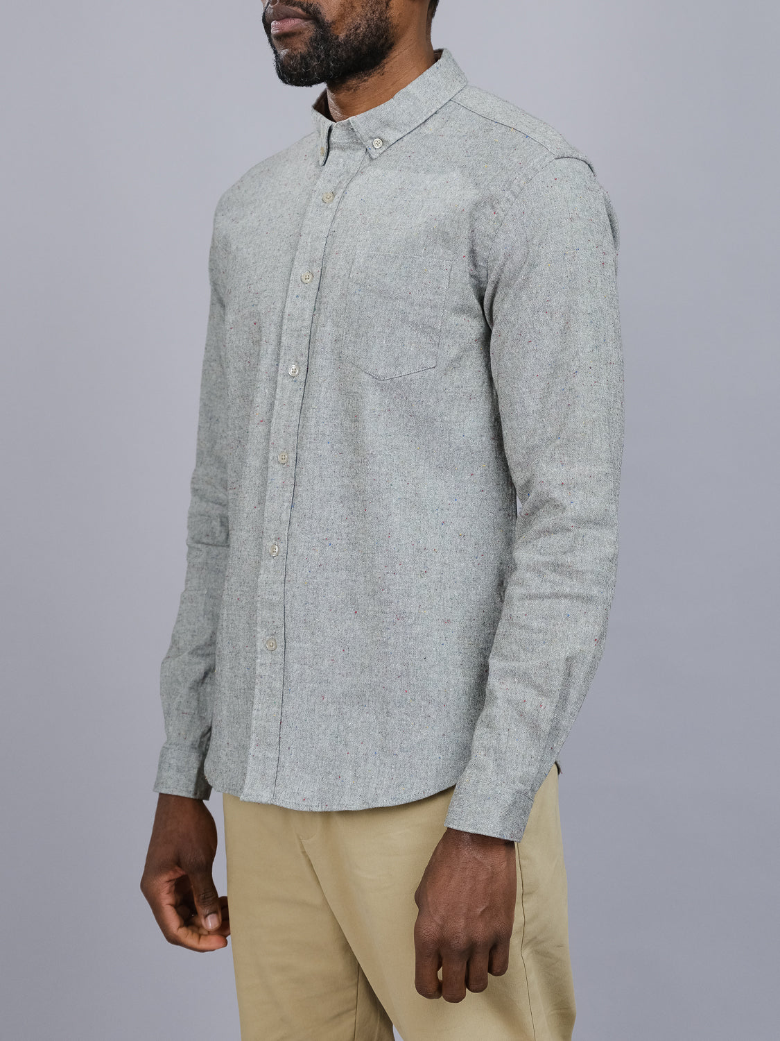 Someone wearing an ID Menswear Allswell Speckled Button Down Shirt, crafted from a premium cotton-linen blend with a subtle pattern, small collar, and chest pocket, pairs it with beige pants against a plain gray backdrop. The person’s head is not visible.