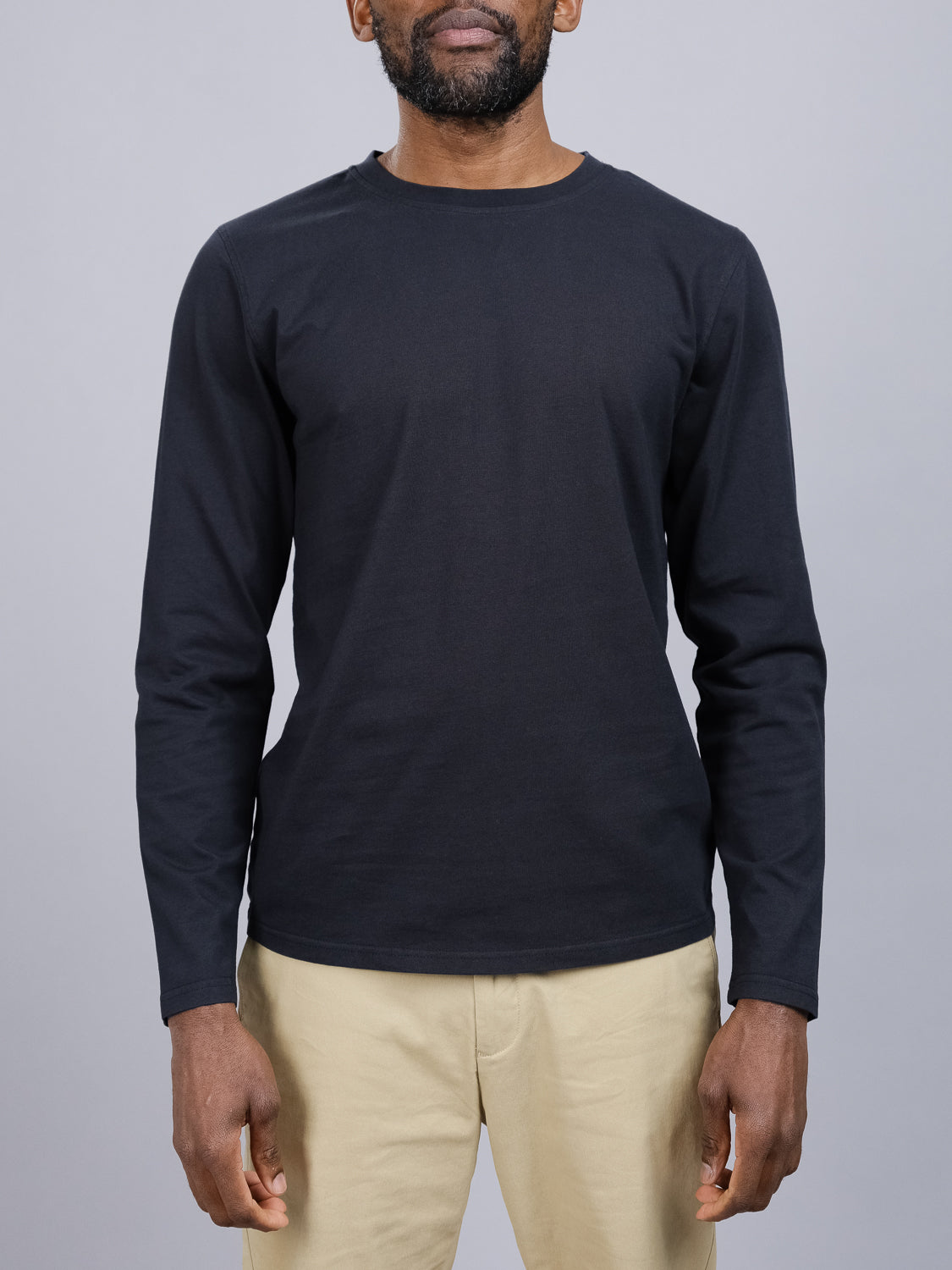 A person dons the Sublime Long Sleeve Tee from ID Menswear, paired with beige pants, standing against a gray background. Their arms are relaxed by their sides in casual wear, with the face not visible.