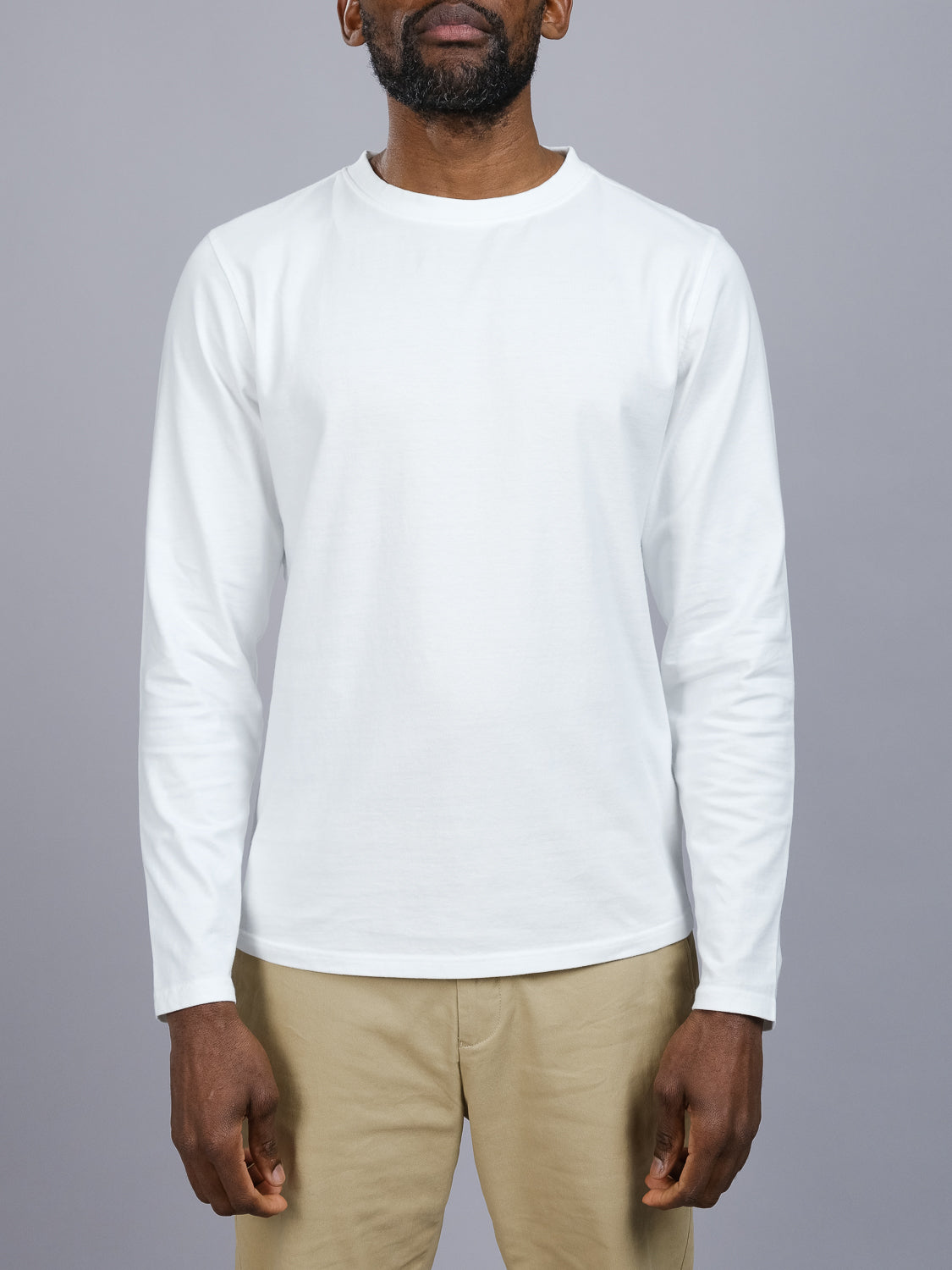 A person models the Sublime Long Sleeve Tee by ID Menswear, a casual organic cotton shirt. Their arms are relaxed by their sides, and theyre paired with beige pants against a gray background, centering on the upper body and clothing.