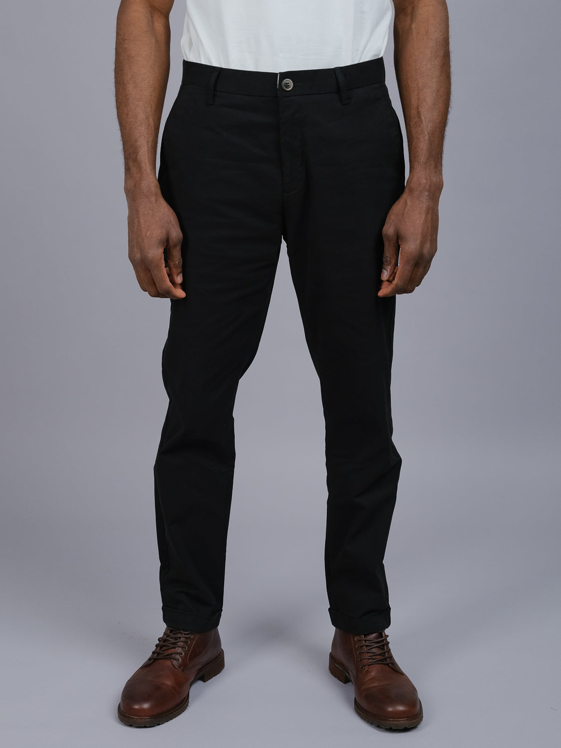 A person in a white shirt, ID Menswears dark Java Chino Pant, and brown leather shoes stands against a gray backdrop. The focus is on the relaxed fit of the pants and the polished footwear.
