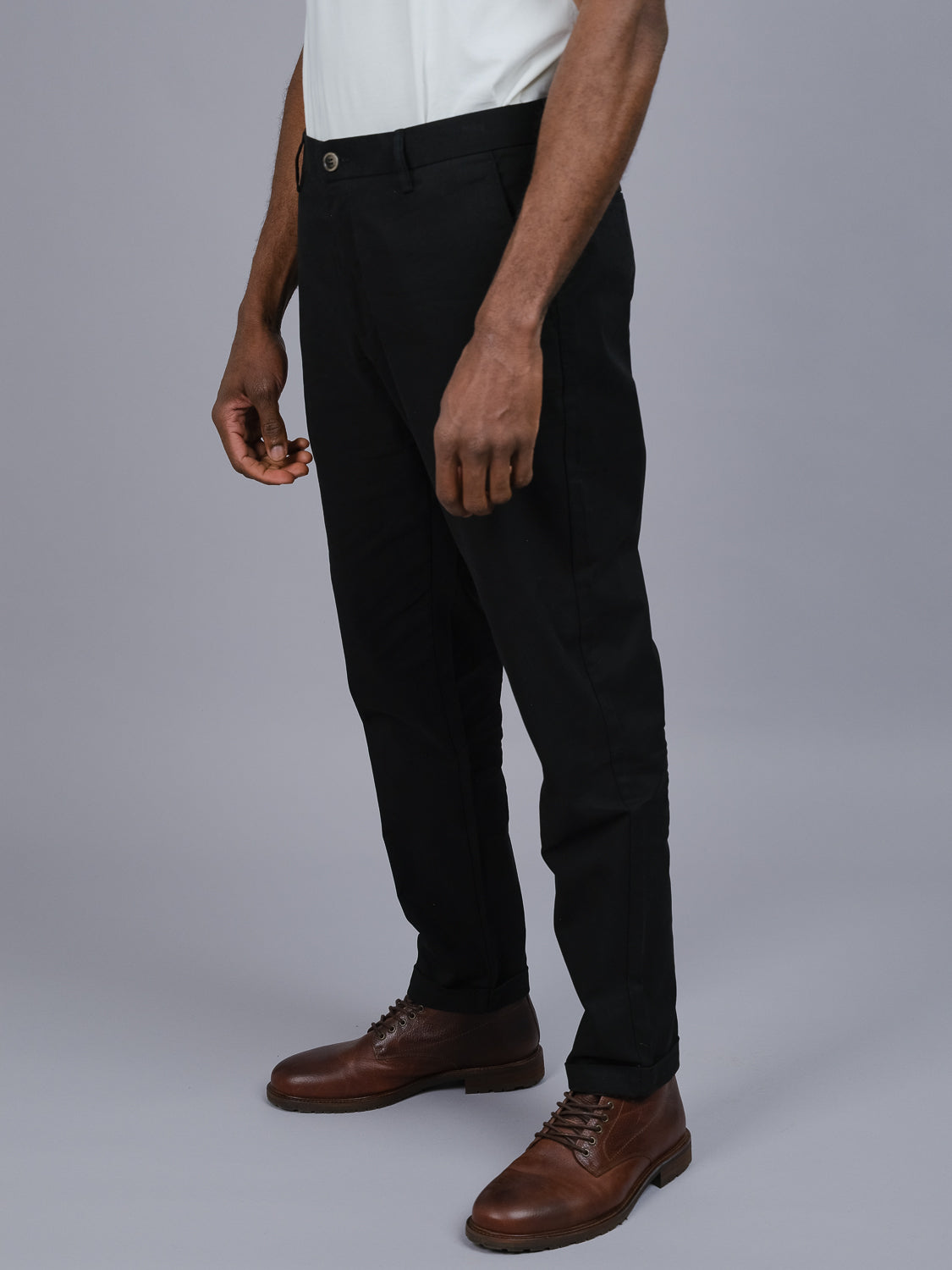 A person in a white shirt and black pants stands against a gray background. The focus is on the lower half, showcasing brown lace-up shoes and relaxed hands. The Java Chino Pants by ID Menswear highlight comfort with their classic cotton twill-inspired fit.