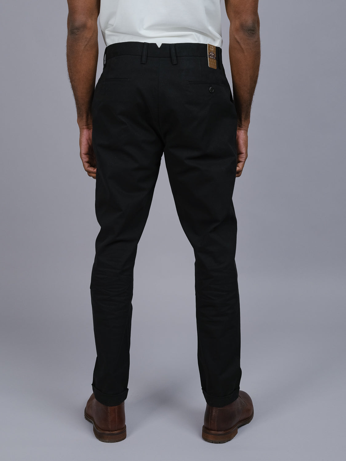 A person is seen from behind, wearing ID Menswears Java Chino Pant in a relaxed fit with pockets and a logo tag, paired with brown shoes and a white shirt. They stand against a gray backdrop, exuding effortless style and sophistication.