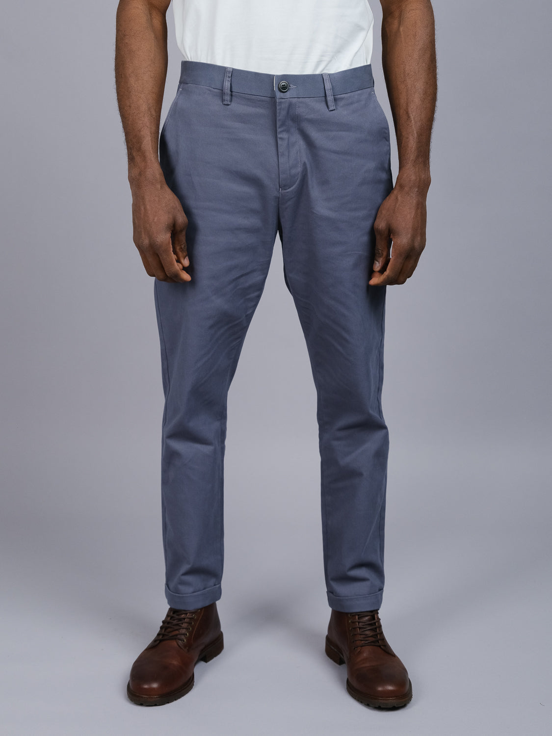In a neutral gray setting, a model dons ID Menswears Java Chino Pants with dark brown leather shoes and a white shirt. The focus is on the relaxed-fit cotton twill trousers and stylish footwear.