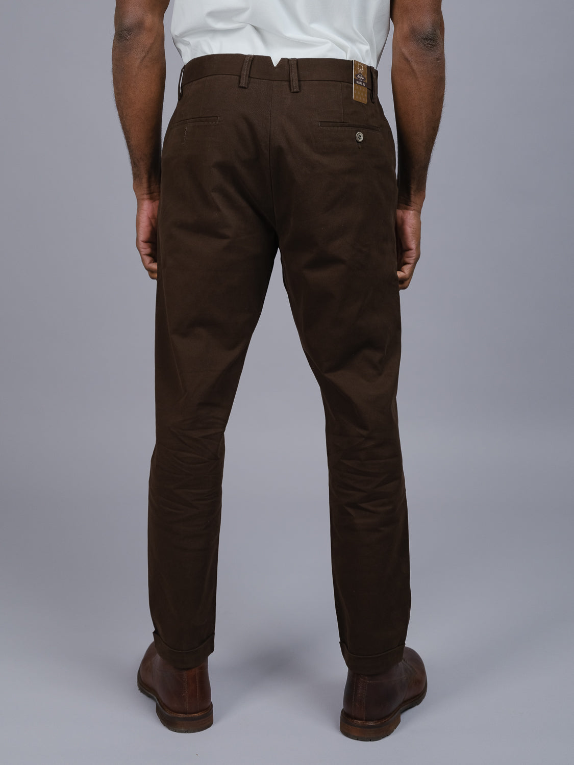 Rear view of a person wearing ID Menswears Java Chino Pant, a relaxed fit dark brown cotton twill trouser with side and back pockets, paired with a white shirt and brown leather shoes, standing against a plain gray background.