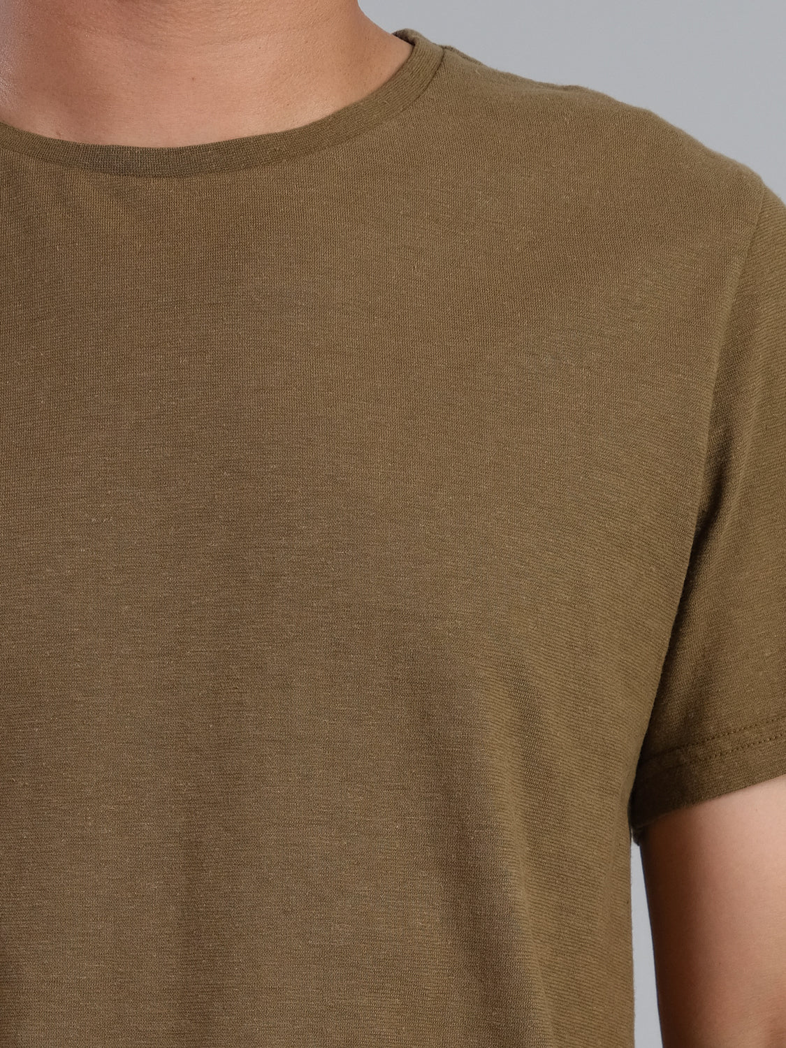 A close-up reveals a person in the Dope Hemp Organic Cotton Tee by ID Menswear, featuring a plain olive green design. The shot captures the neck and upper torso against a neutral backdrop, showcasing its sustainable fabric.