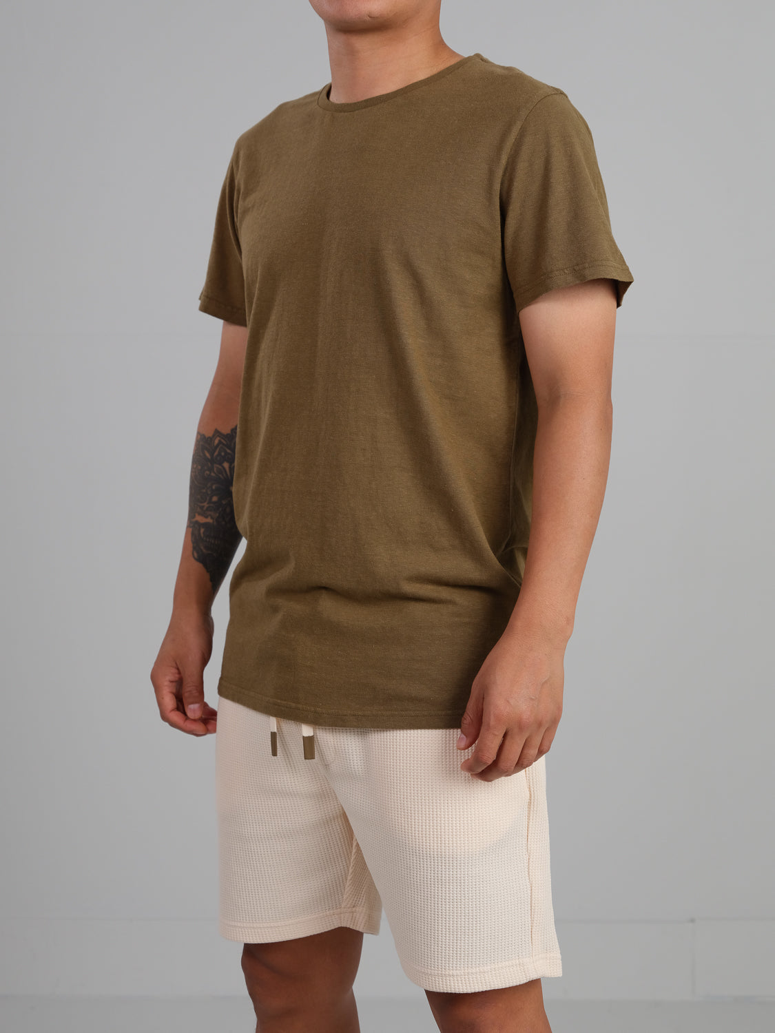 A person in a Dope Hemp Organic Cotton Tee from ID Menswear and beige shorts made from sustainable fabric stands against a gray background. A tattoo on their right forearm is visible, with focus on the eco-friendly materials of the torso and legs.