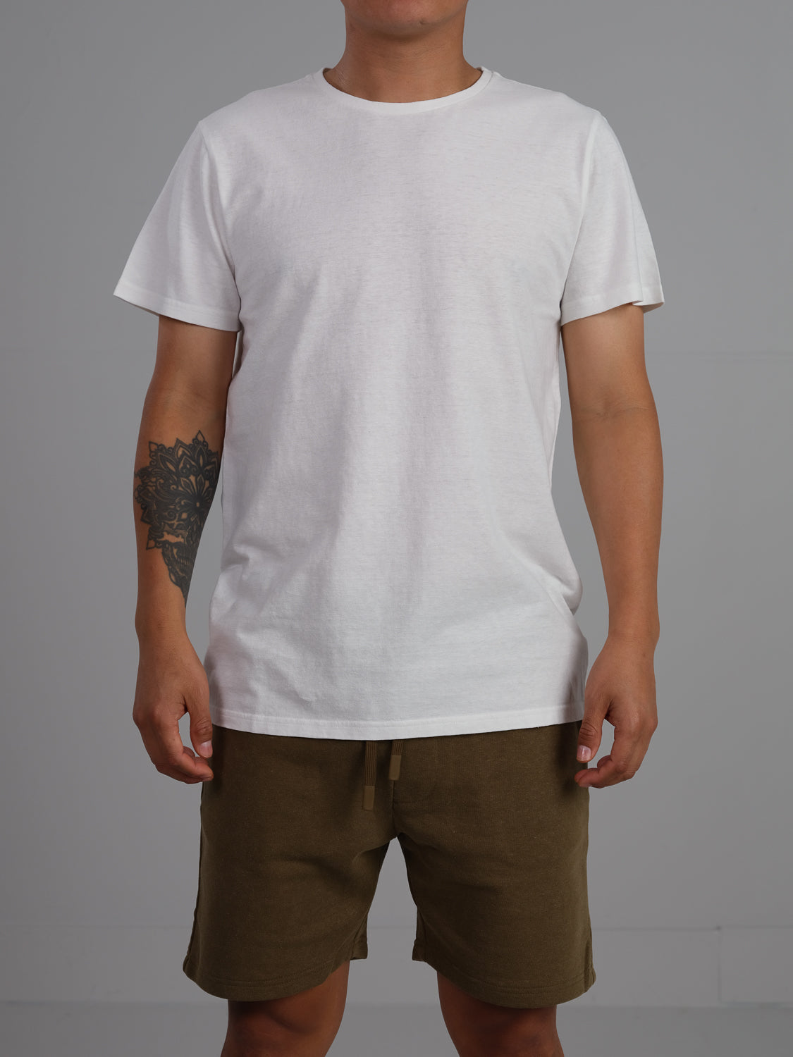 A person in a Dope Hemp Organic Cotton Tee by ID Menswear and olive shorts stands against a neutral backdrop, their arms relaxed with an intricate forearm tattoo visible.