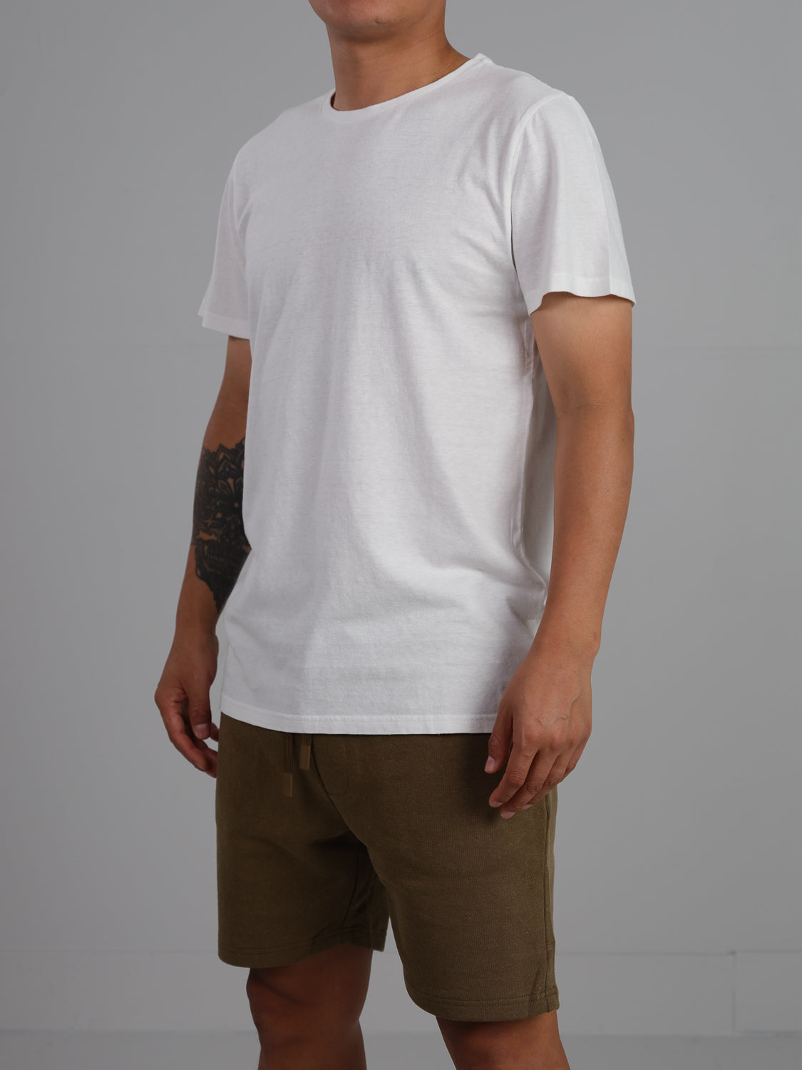 A person in an ID Menswear Dope Hemp Organic Cotton Tee and olive green hemp cotton shorts stands against a gray backdrop. They have a forearm tattoo, and the image is cropped above the knees, emphasizing the sustainable fabric choice.