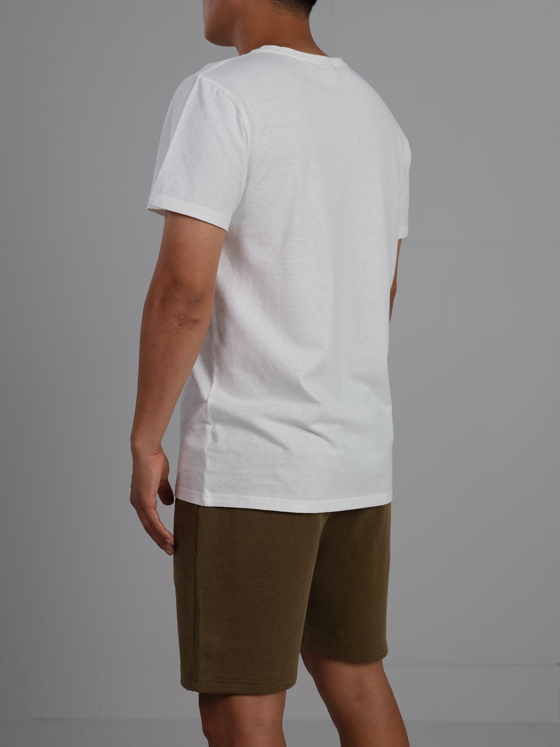 Facing away against a gray background, a person showcases the back of an ID Menswear Dope Hemp Organic Cotton Tee in white, paired with brown shorts.