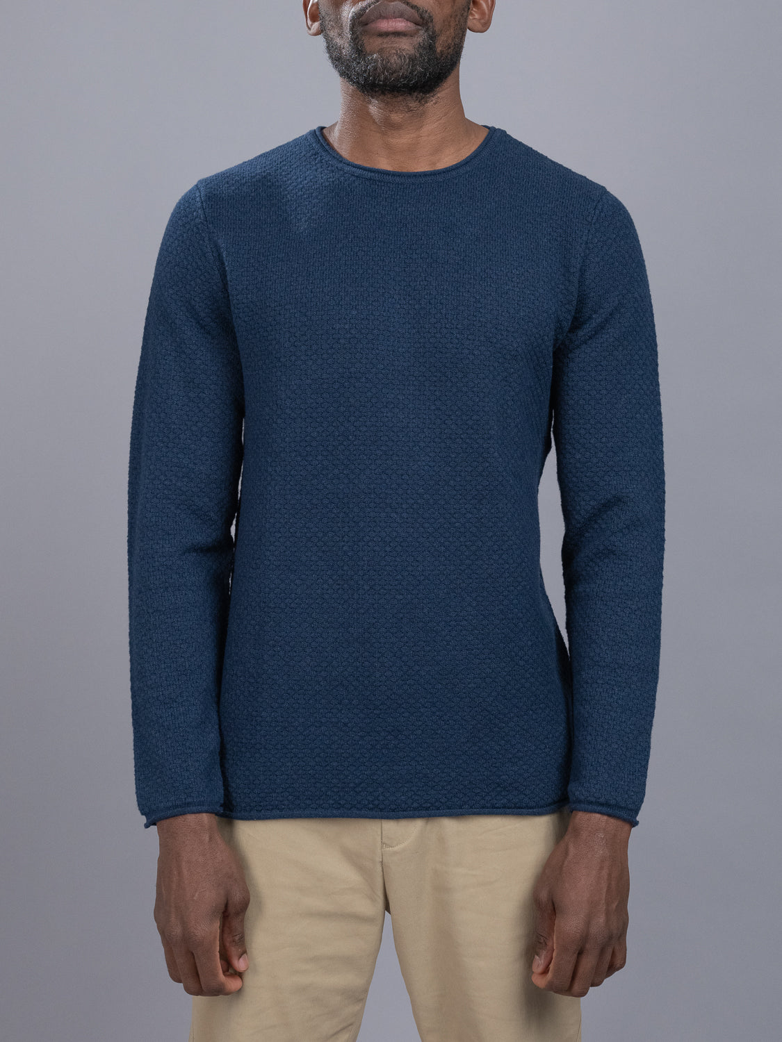 A person is wearing a textured blue Westside Rolled Crew Neck Sweater from ID Menswear and beige pants, standing against a plain gray background. The persons face is not visible in the image.