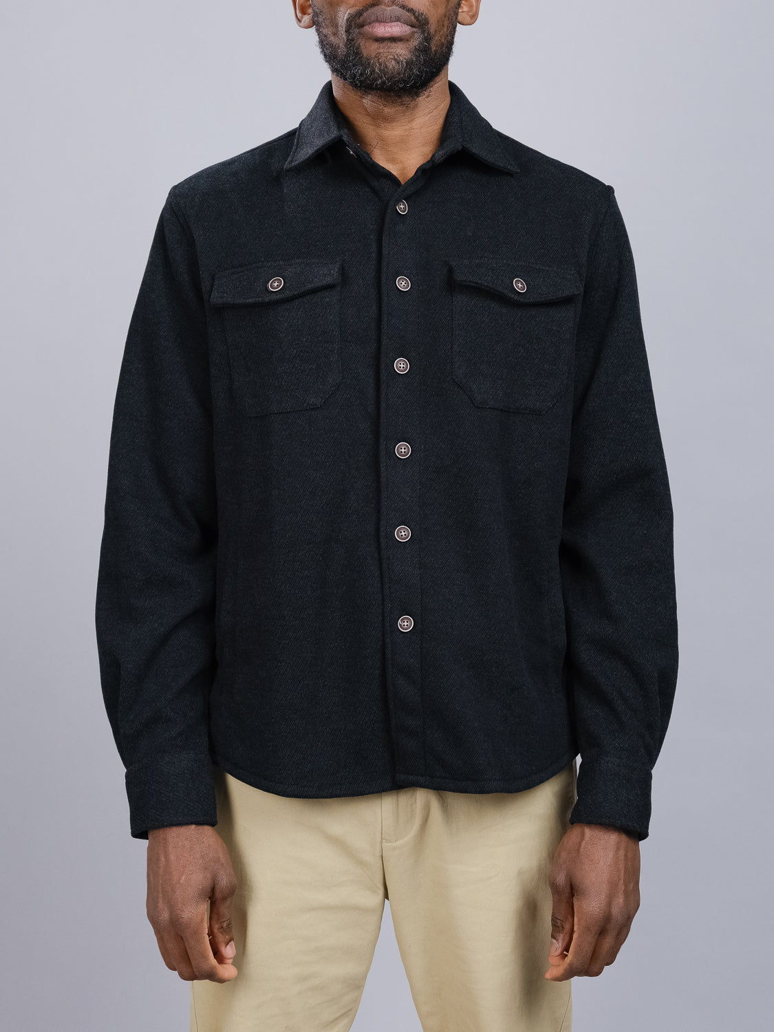 Someone is seen against a gray backdrop, wearing an ID Menswear black Yukon Shirt Jacket with buttoned cuffs and a pointed collar. Ideal for transitional weather and casual outings, it features two front pockets and pairs well with beige pants, though their face remains unseen.