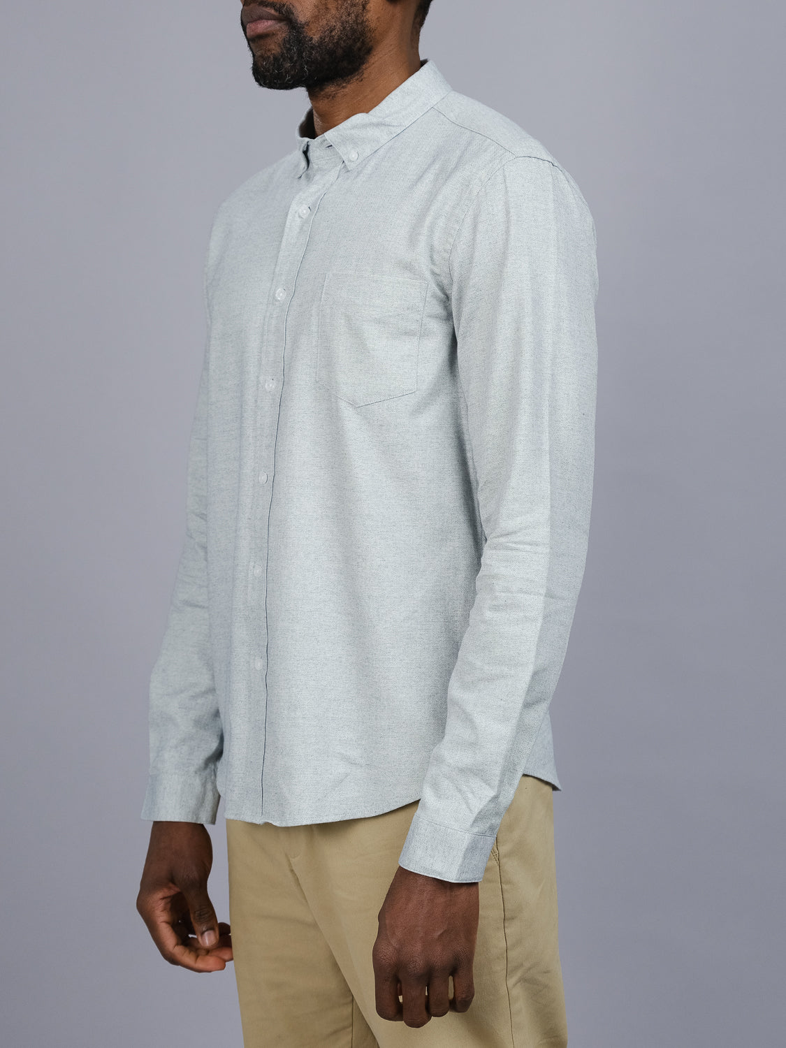 A person in a light gray Maya Fine Herringbone Shirt by ID Menswear, featuring a chest pocket and made of 100% cotton, along with tan pants, stands sideways against a plain gray background. Their face is not visible in the image.