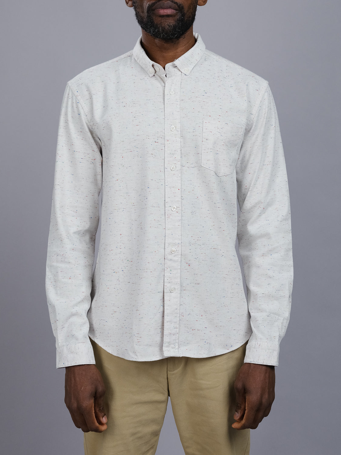 Someone wearing the Allswell Speckled Button Down Shirt from ID Menswear, made with a cotton-linen blend and matched with beige pants. The shirts colored speckles subtly stand out against its plain gray background, highlighting its versatile design.