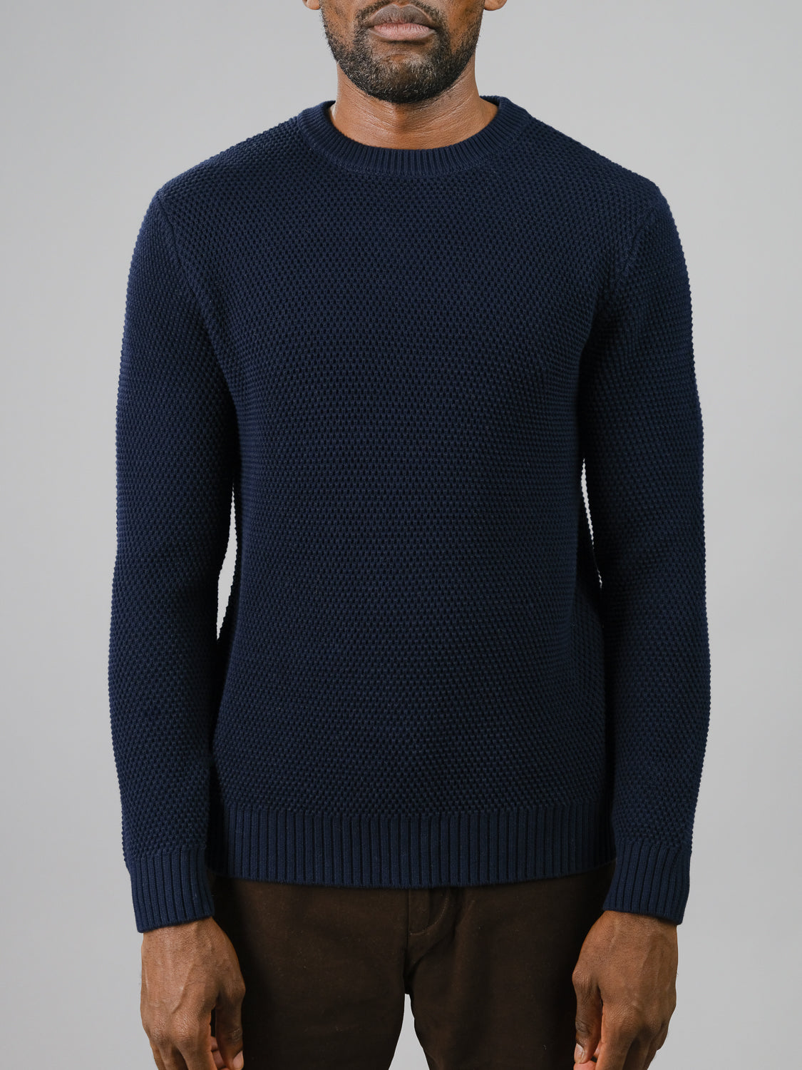 An individual models a Quebec Chunky Sweater by ID Menswear, featuring a cozy navy blue Moss knit design, paired with brown pants. They stand against a plain gray backdrop with their face hidden.