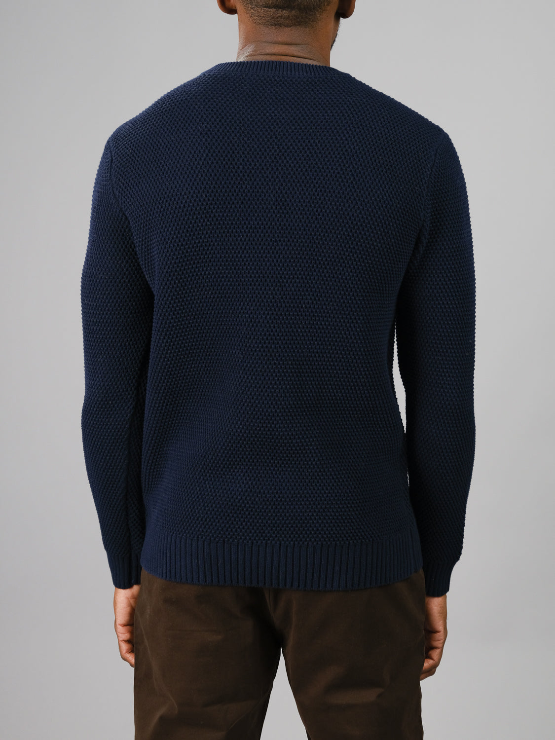A person is seen from behind wearing a dark blue Quebec Chunky Sweater by ID Menswear, featuring a cozy Moss knit pattern. They pair it with dark brown pants against a plain gray background.