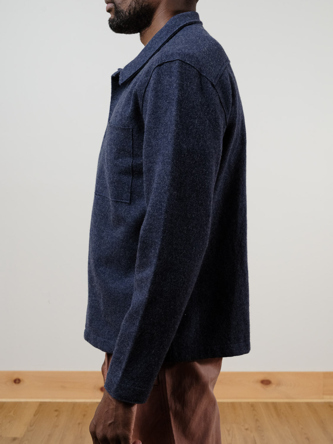 A man stands sideways, showing his left side, wearing the Marine Layer Mendocino Wool Overshirt in navy blue and brown pants. The setting features a plain light wall with a wooden floor and baseboard.