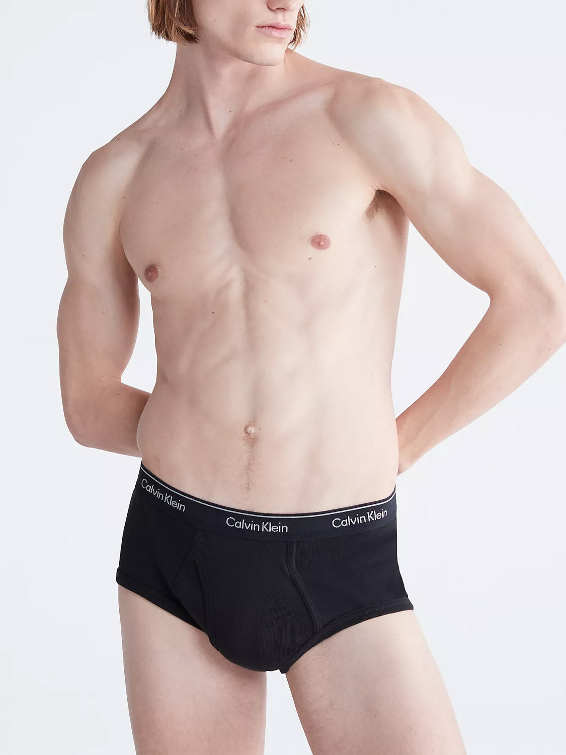 A shirtless person stands with hands on hips, wearing Calvin Klein NB3999 3-Pack Classic Cotton Briefs in black. The image highlights the torso and upper legs against a plain background.