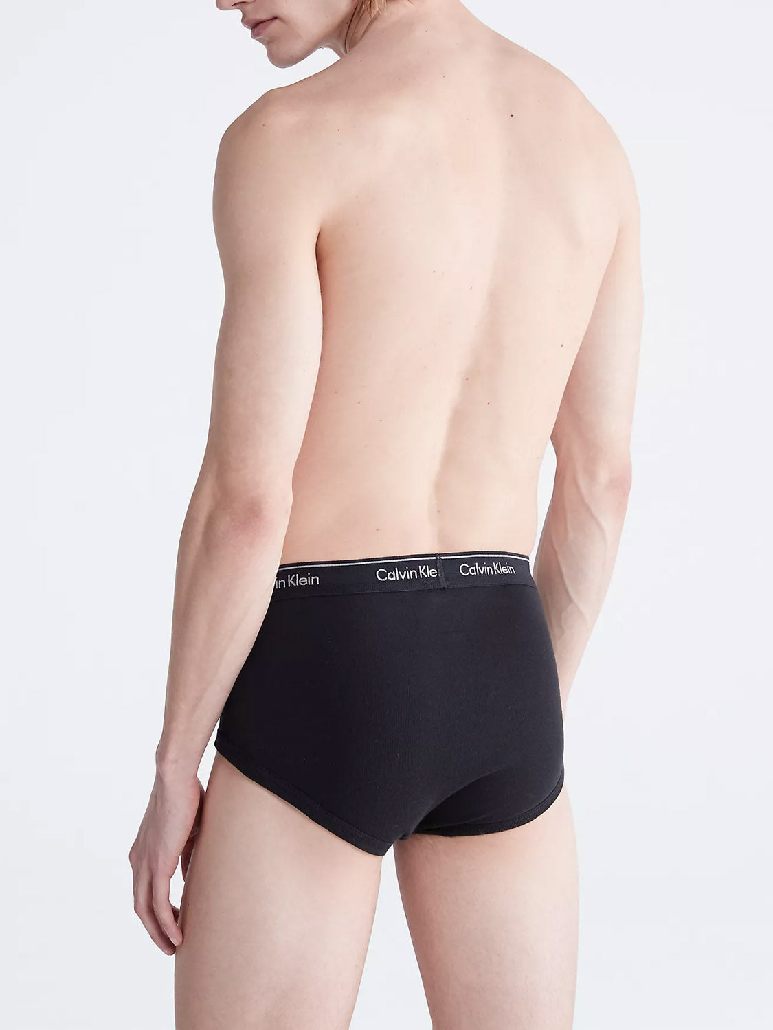 A shirtless person is shown from the back wearing Calvin Klein NB3999 3-Pack Classic Cotton Briefs in black, featuring a classic fit and a white-logo waistband. The background is plain and light-colored.