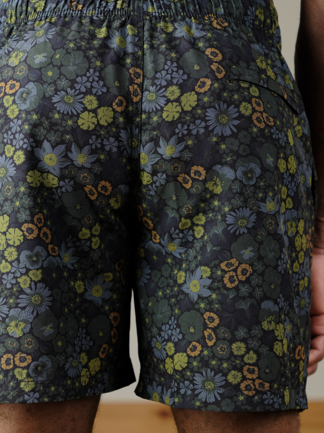 A close-up of a person wearing ID Menswear’s Wild Flower Fast Dry 4-Way Stretch Swim Trunk, featuring a colorful floral pattern in green, blue, and yellow; the image focuses on the back of the shorts.