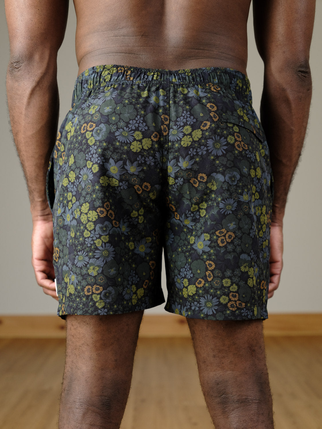 A person stands indoors wearing ID Menswear’s Wild Flower Fast Dry 4-Way Stretch Swim Trunk, featuring a dark base with green, yellow, and blue floral prints. The background is softly blurred with a light wall and wooden floor.