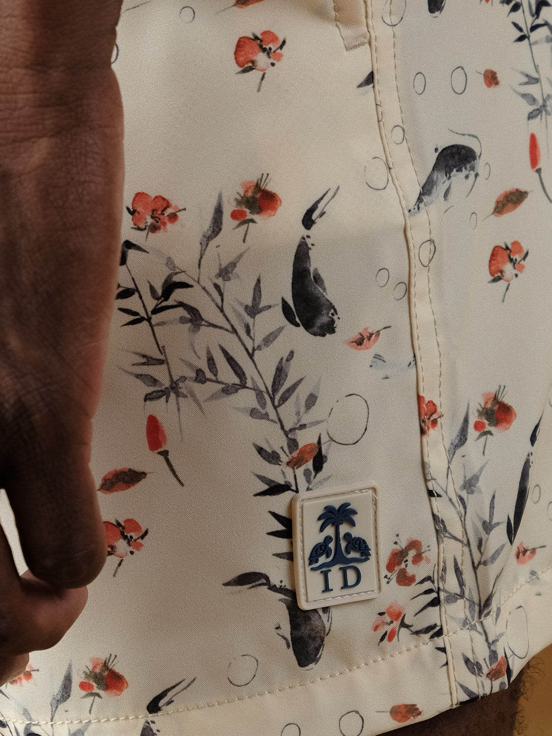 Close-up of Koi Fast Dry 4-Way Stretch Swim Trunk by ID Menswear in cream, featuring a floral and wildlife print with red flowers, leaves, small animals, and a rectangular “ID” palm tree patch near the hem.