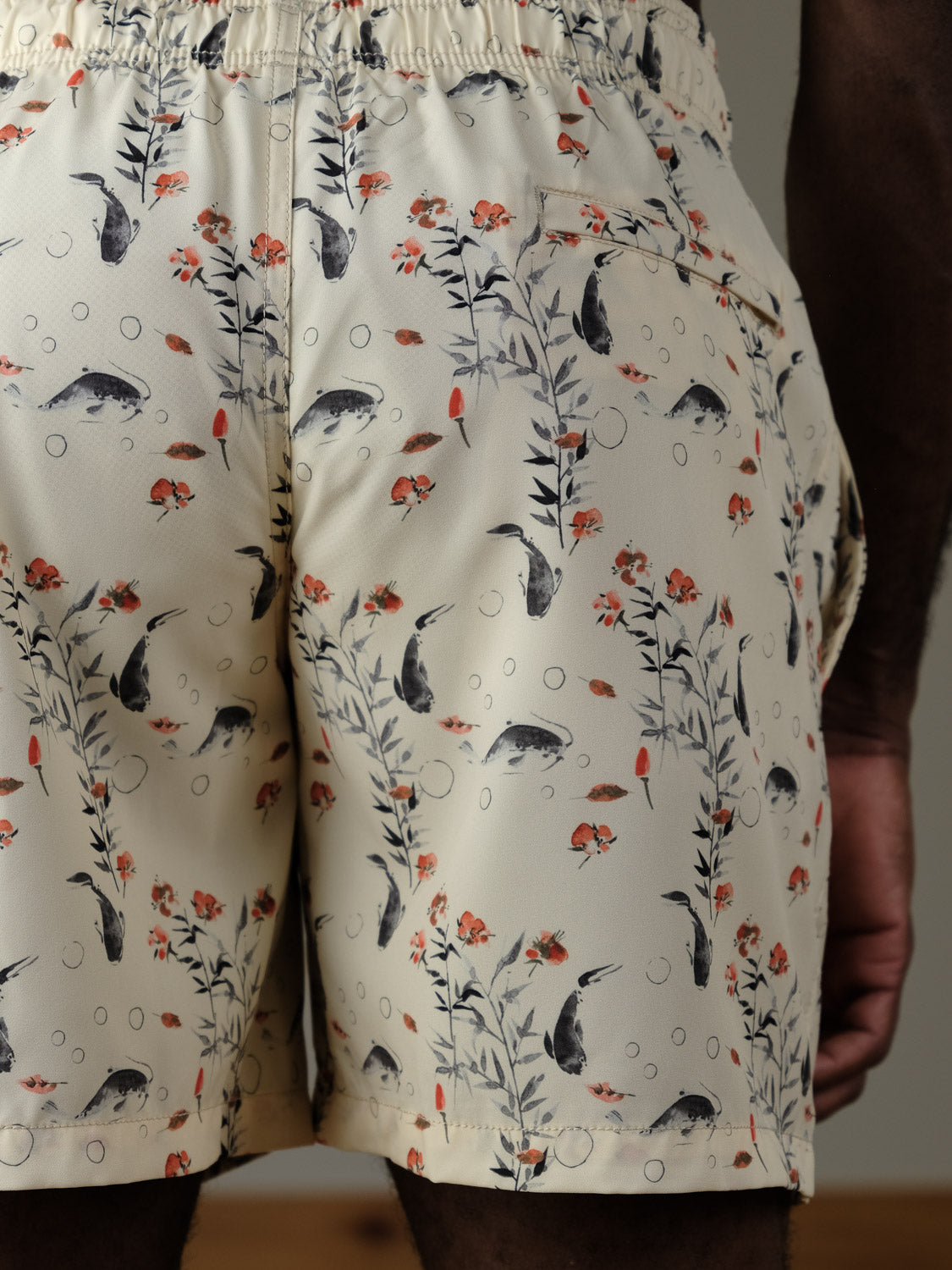 A close-up of the back of a person wearing the Koi Fast Dry 4-Way Stretch Swim Trunk by ID Menswear, featuring a cream base with playful fish, plants, bubbles, and orange flowers. These quick-dry shorts have an elastic waist and back pocket—ideal for water adventures.