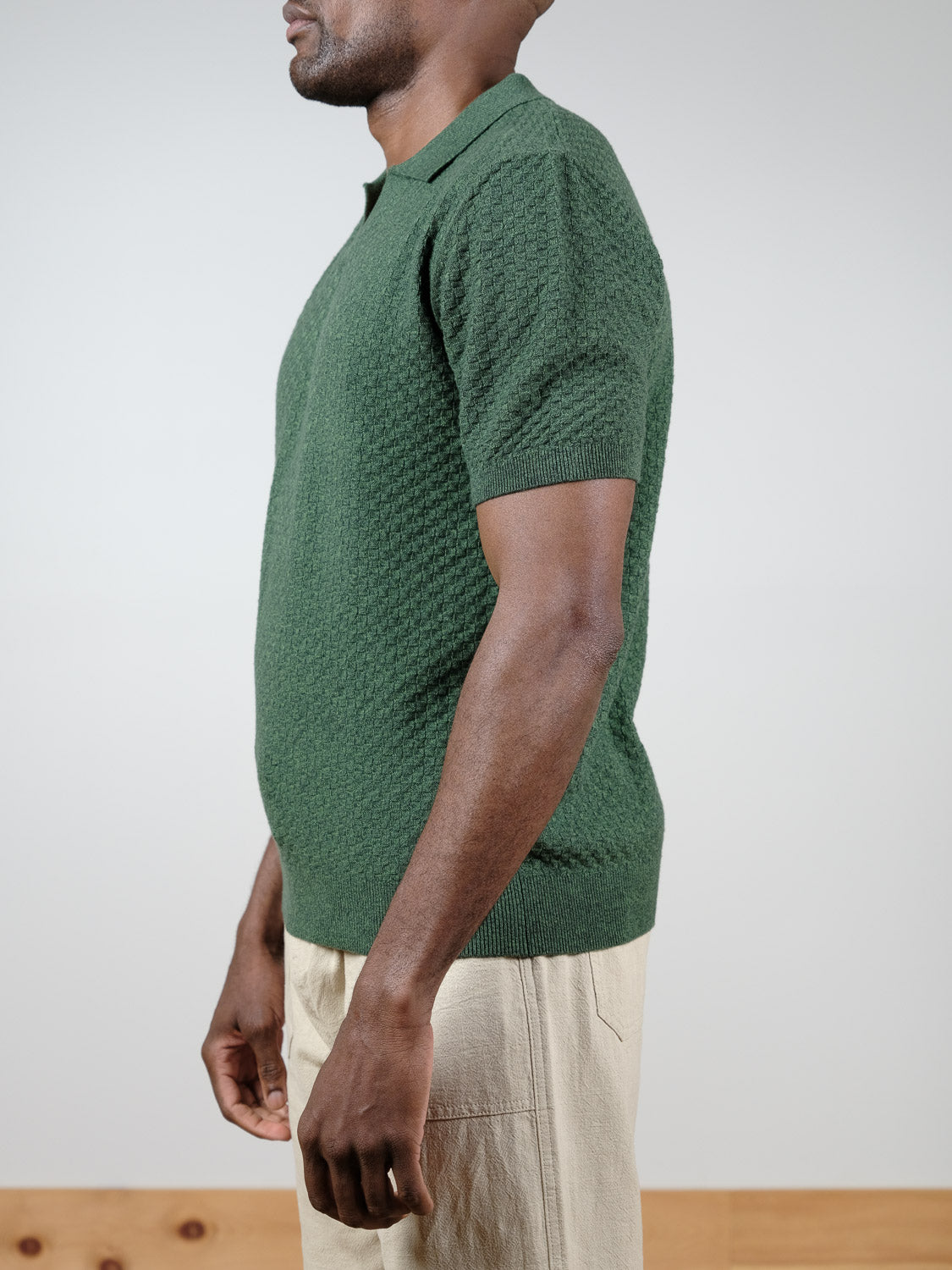 A man stands sideways in a textured green Cairo Knitted Cotton Polo from ID Menswear and light beige pants against a plain light background.