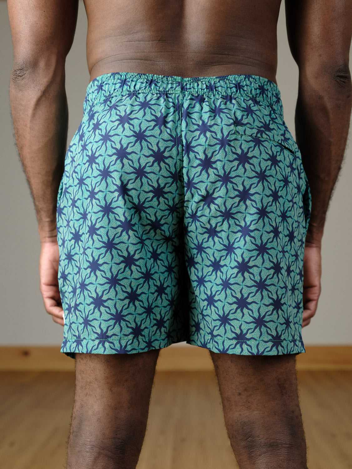 A person stands indoors facing away from the camera, wearing the Arica Fast Dry 4-Way Stretch Swim Trunk by ID Menswear in teal and navy with an elastic waistband. The setting features neutral tones and wooden flooring.
