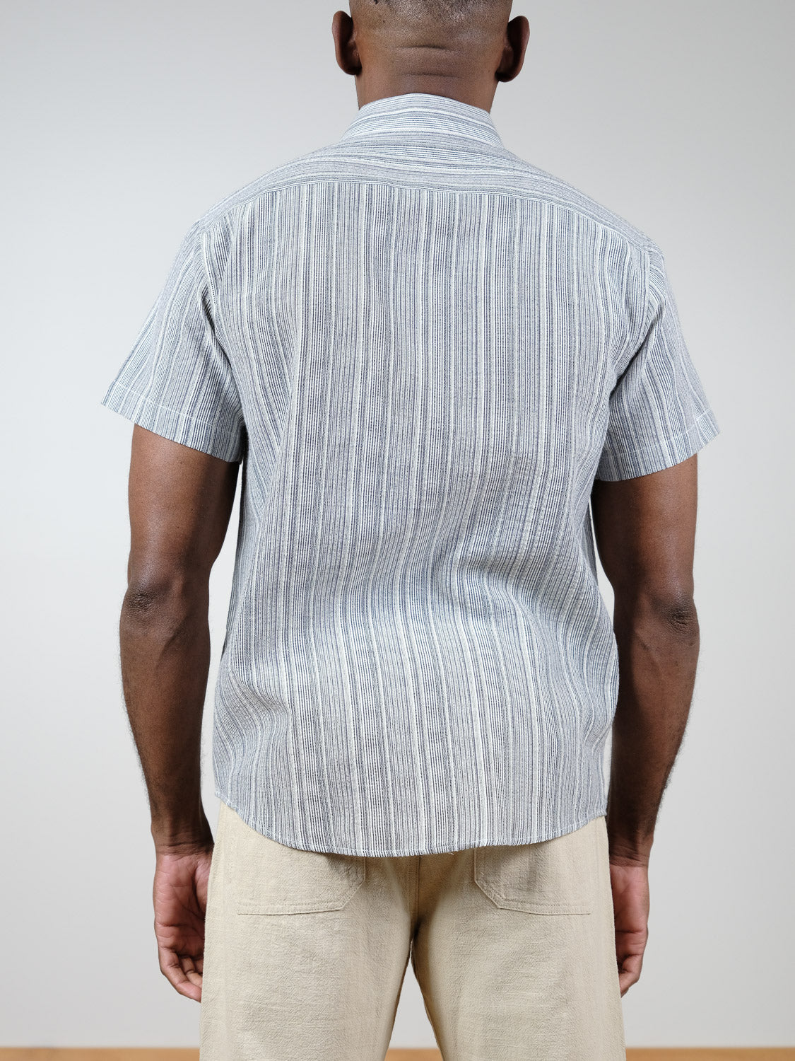 A person faces away from the camera wearing the ID Menswear Strider Short Sleeve Shirt in light gray and white stripes with beige pants, set against a plain light background.