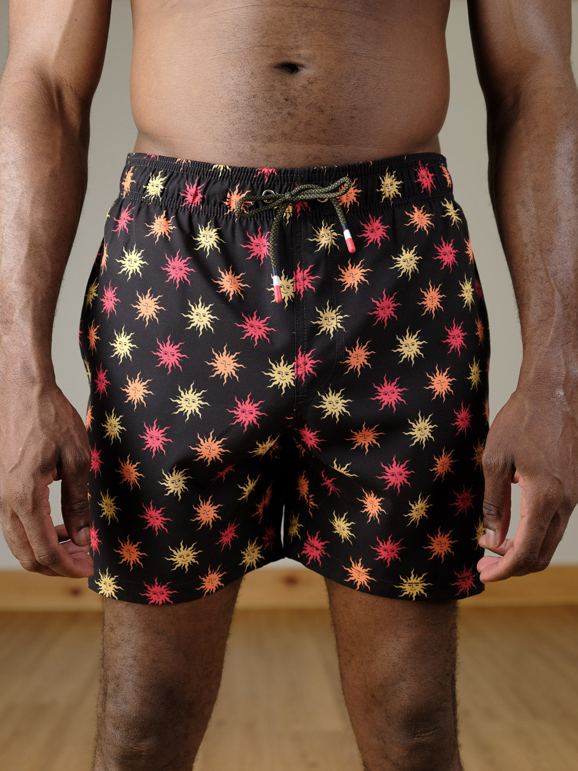 A person stands indoors on a wooden floor wearing ID Menswear’s Sunshine Fast Dry 4-Way Stretch Swim Trunk, featuring a black base with a colorful red, yellow, and orange starburst pattern.
