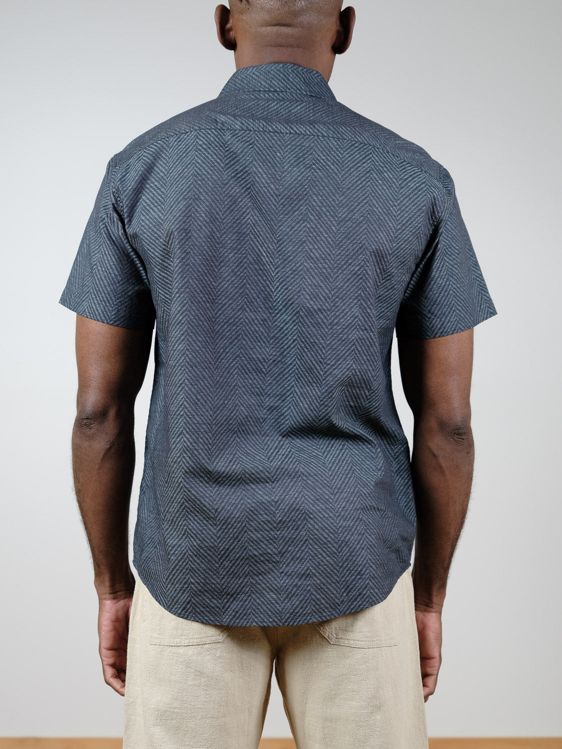 A person is shown from the back wearing the ID Menswear Reveller Short Sleeve Shirt, featuring a relaxed fit with a dark geometric weave, paired with light-colored pants against a plain light background.