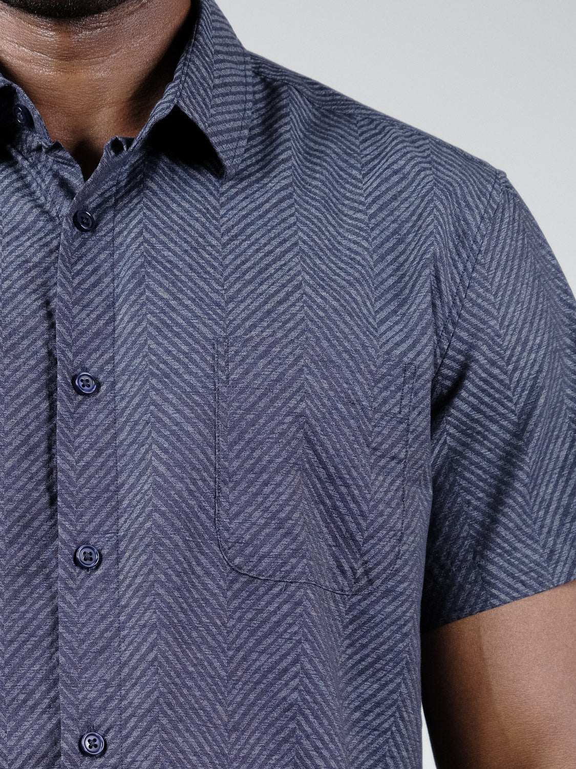 Close-up of a person wearing the ID Menswear Reveller Short Sleeve Shirt in dark blue, featuring a relaxed fit, geometric chevron weave, and left chest pocket. Only the upper torso, part of the neck, and arms are visible.
