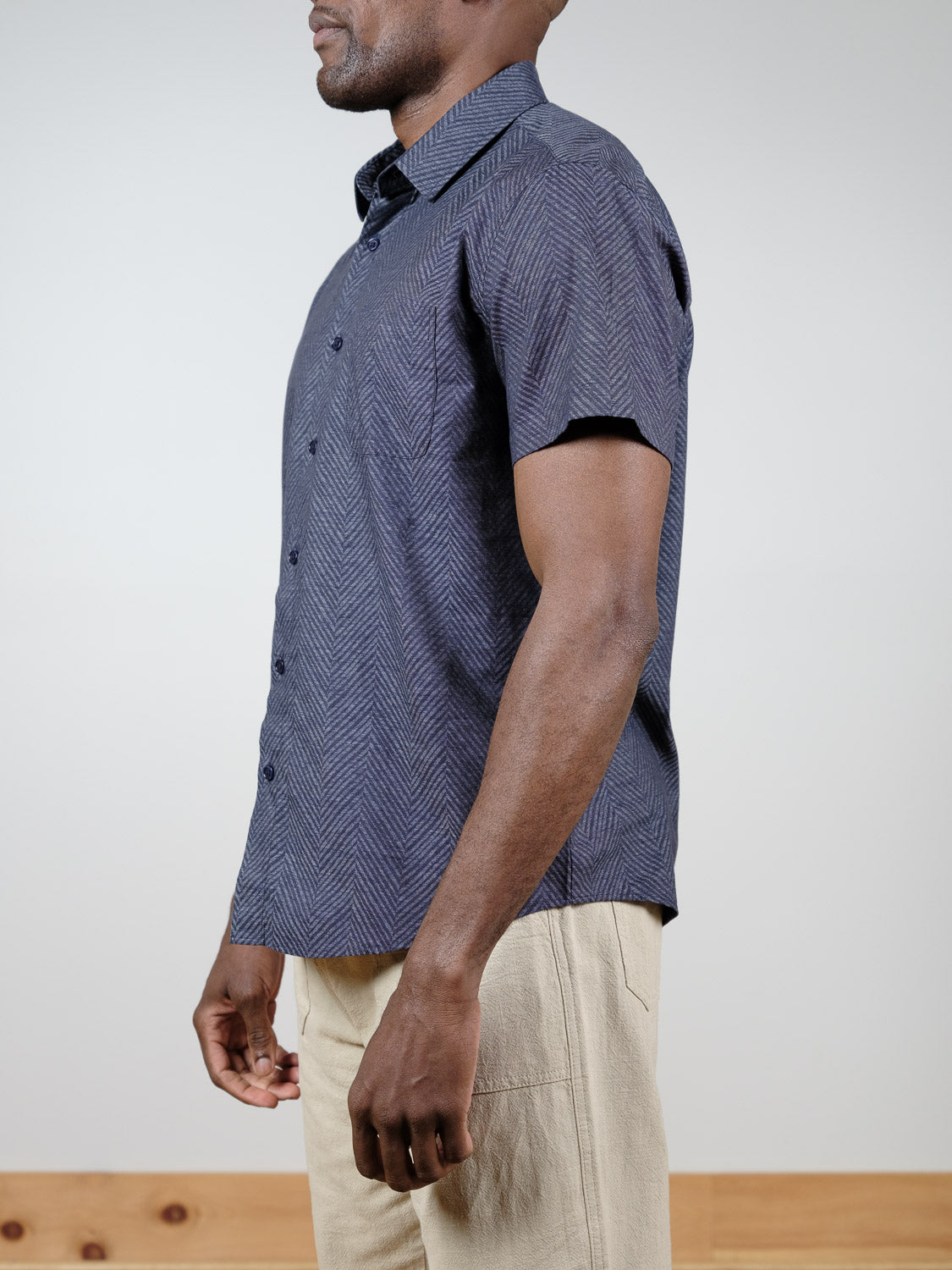 A man in profile wears the Reveller Short Sleeve Shirt by ID Menswear—a relaxed fit, dark blue patterned button-up made from breathable slub cotton—paired with light beige pants against a plain background.